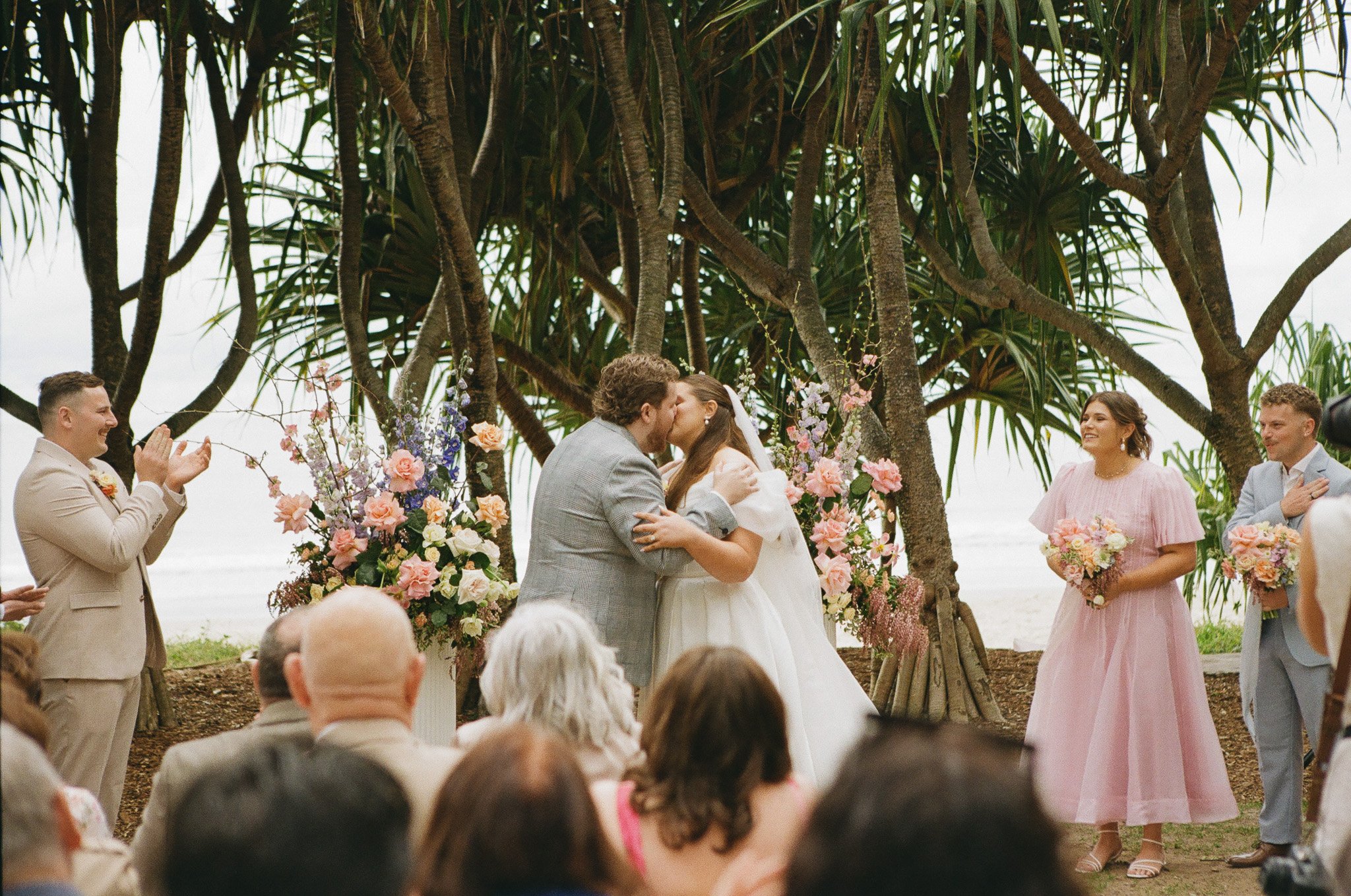 A wedding ceremony outdoors near the beach, with a couple kissing at the altar, surrounded by floral arrangements and greenery. Guests are applauding and standing in front of the couple.