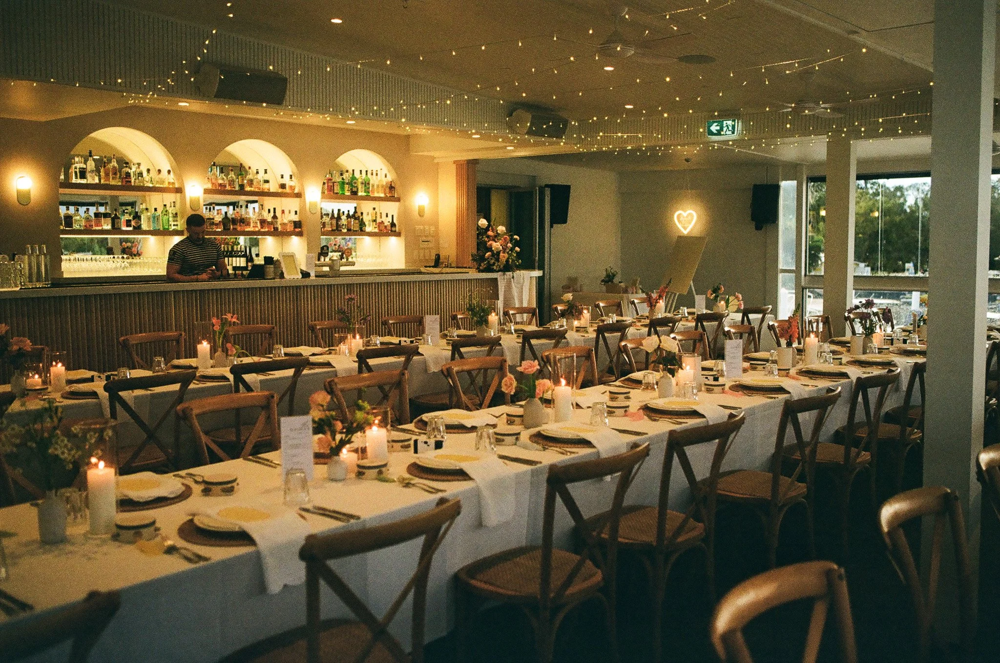 Event space decorated with long tables set for a celebration, with tableware, candles, and floral centerpieces, illuminated by string lights and candles, with a bar in the background and a neon heart sign.