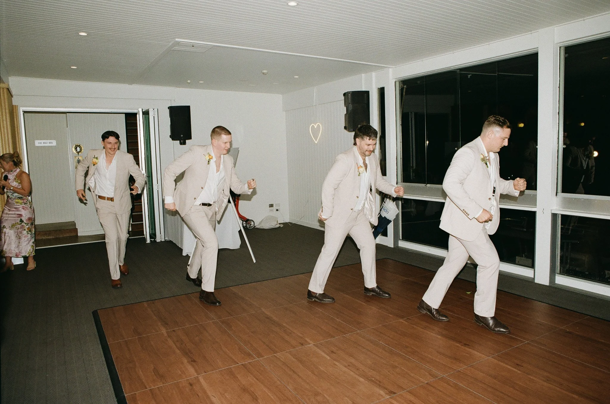 A group of men in light-colored suits dancing on a wooden floor at a wedding reception. They are smiling and appear to be enjoying themselves.