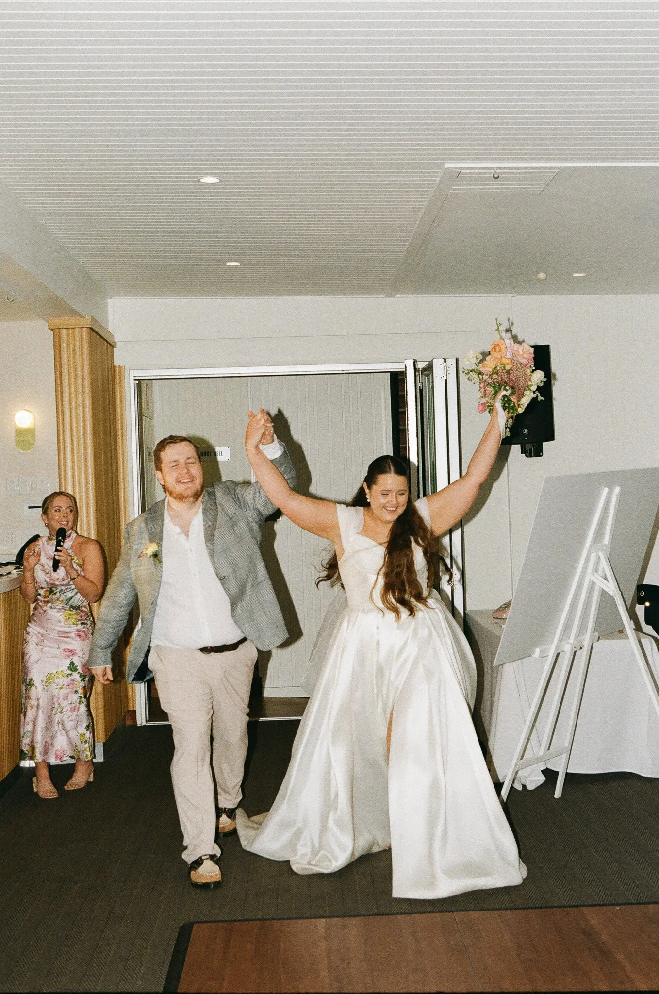 A bride and groom holding hands and celebrating at their wedding reception indoors, with a woman in a floral dress in the background holding a microphone.