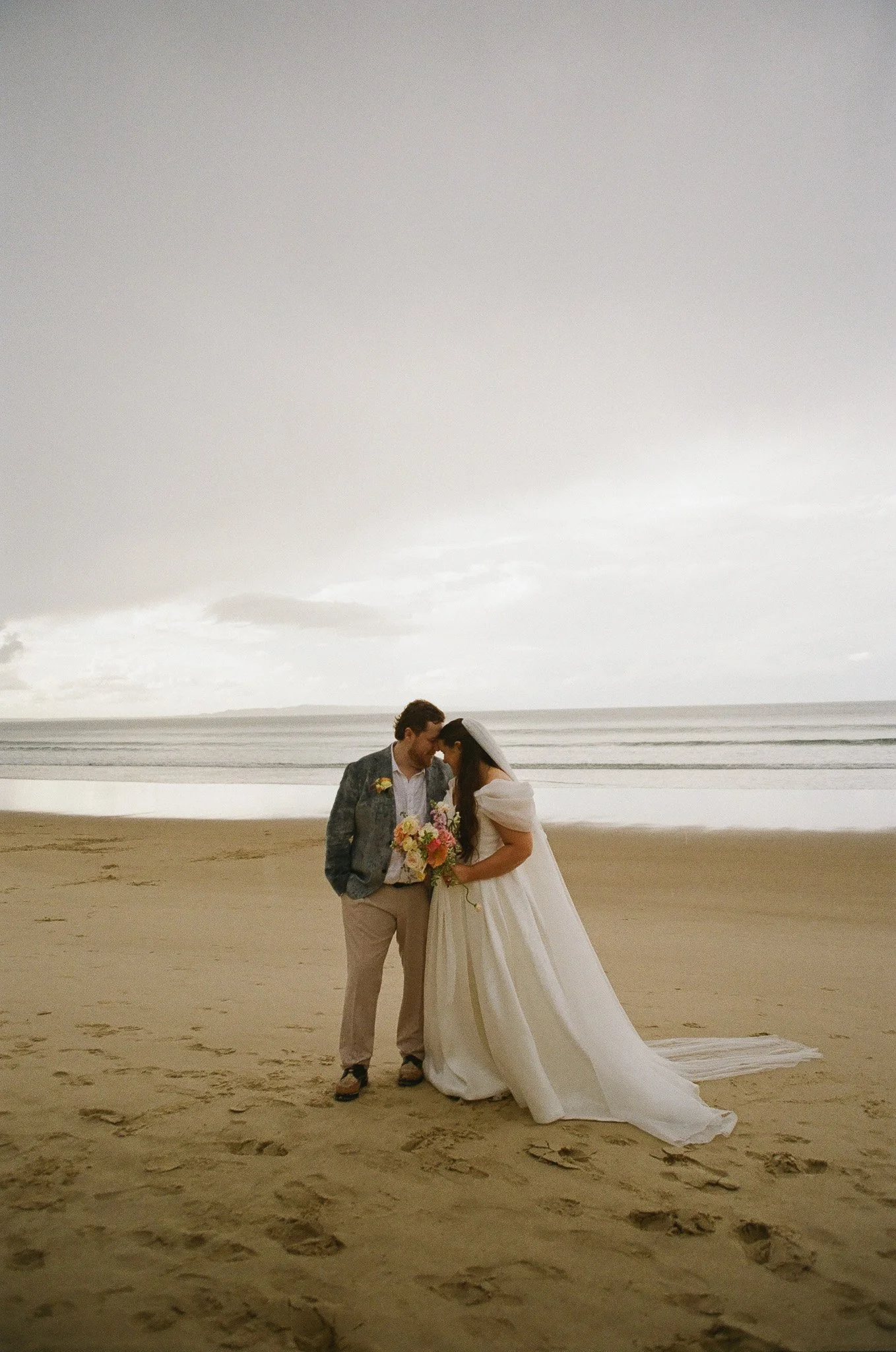 A bride and groom on a beach, standing close with foreheads touching, holding a bouquet of flowers, under a cloudy sky.