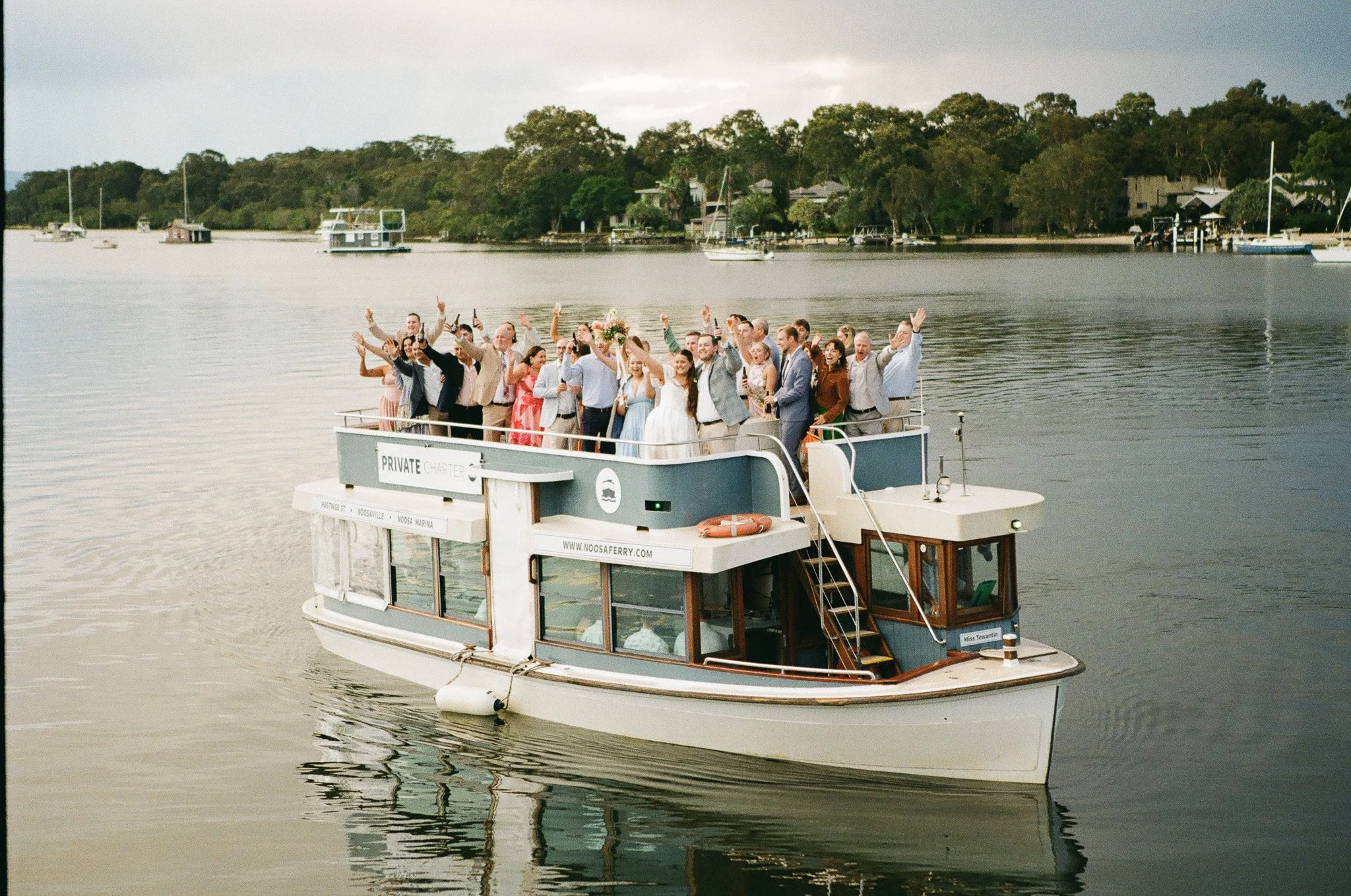 A group of people celebrating on a boat on a calm river, with trees and houses in the background.