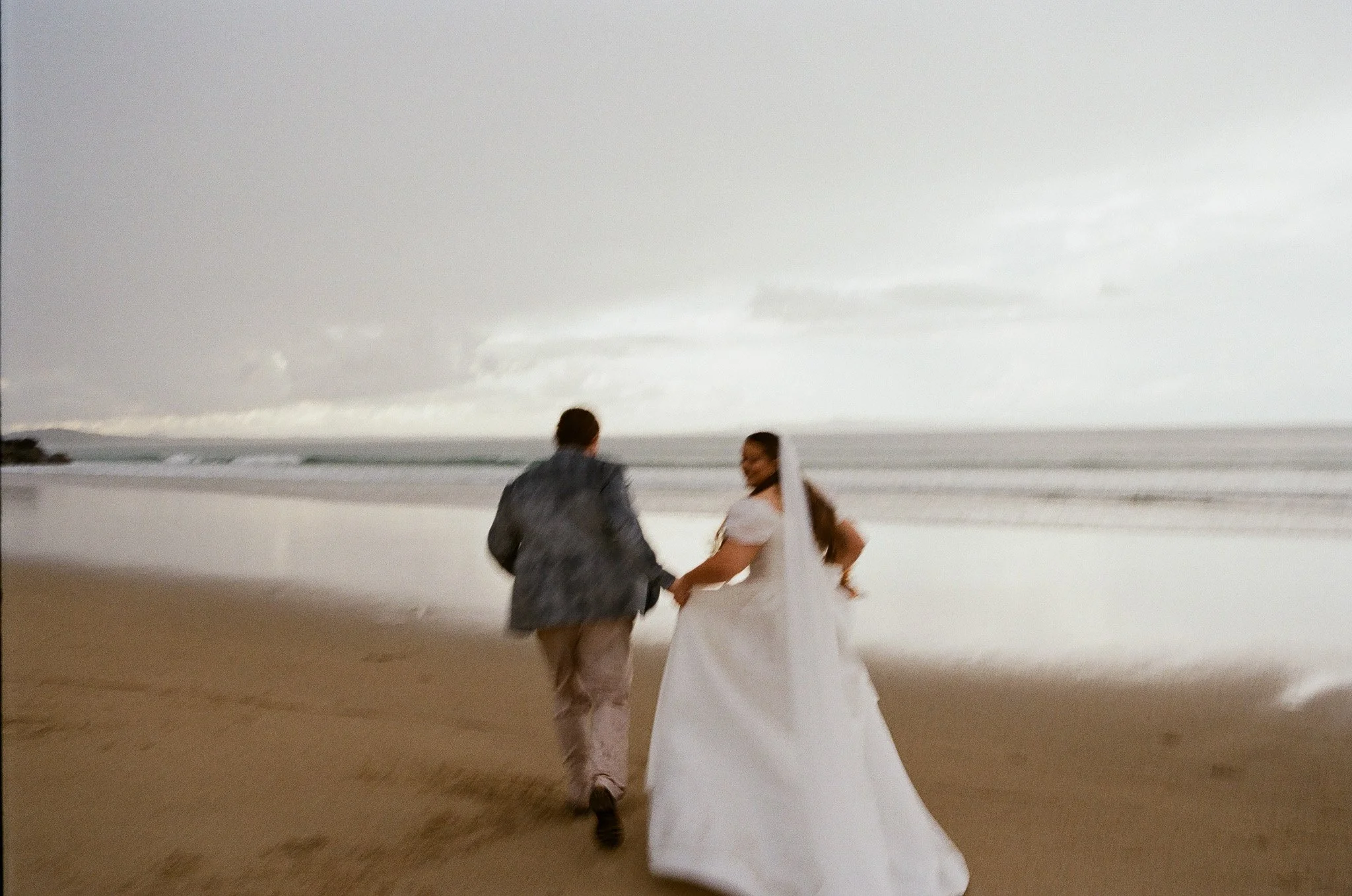 A couple in wedding attire walking along the beach holding hands, with the ocean and sky in the background.