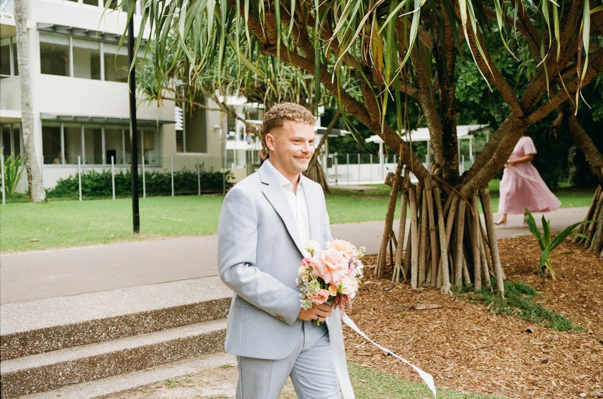 A man in a light gray suit holding a pink and peach flower bouquet, standing outdoors near a large tree with a suburban residential background.