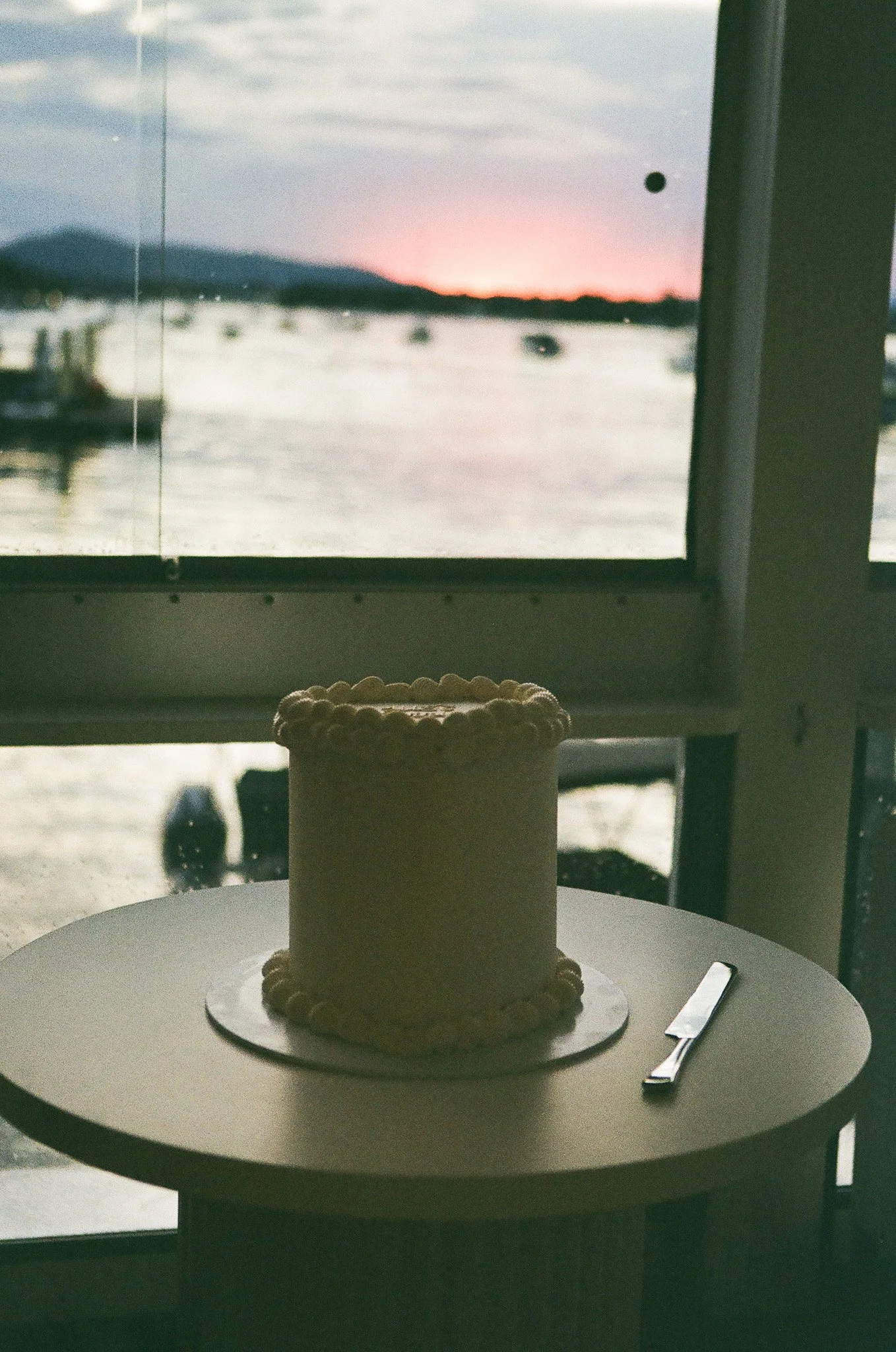 A white cake with decorative piping on a round white table, next to a cake knife, with a window showing a sunset over a body of water in the background.