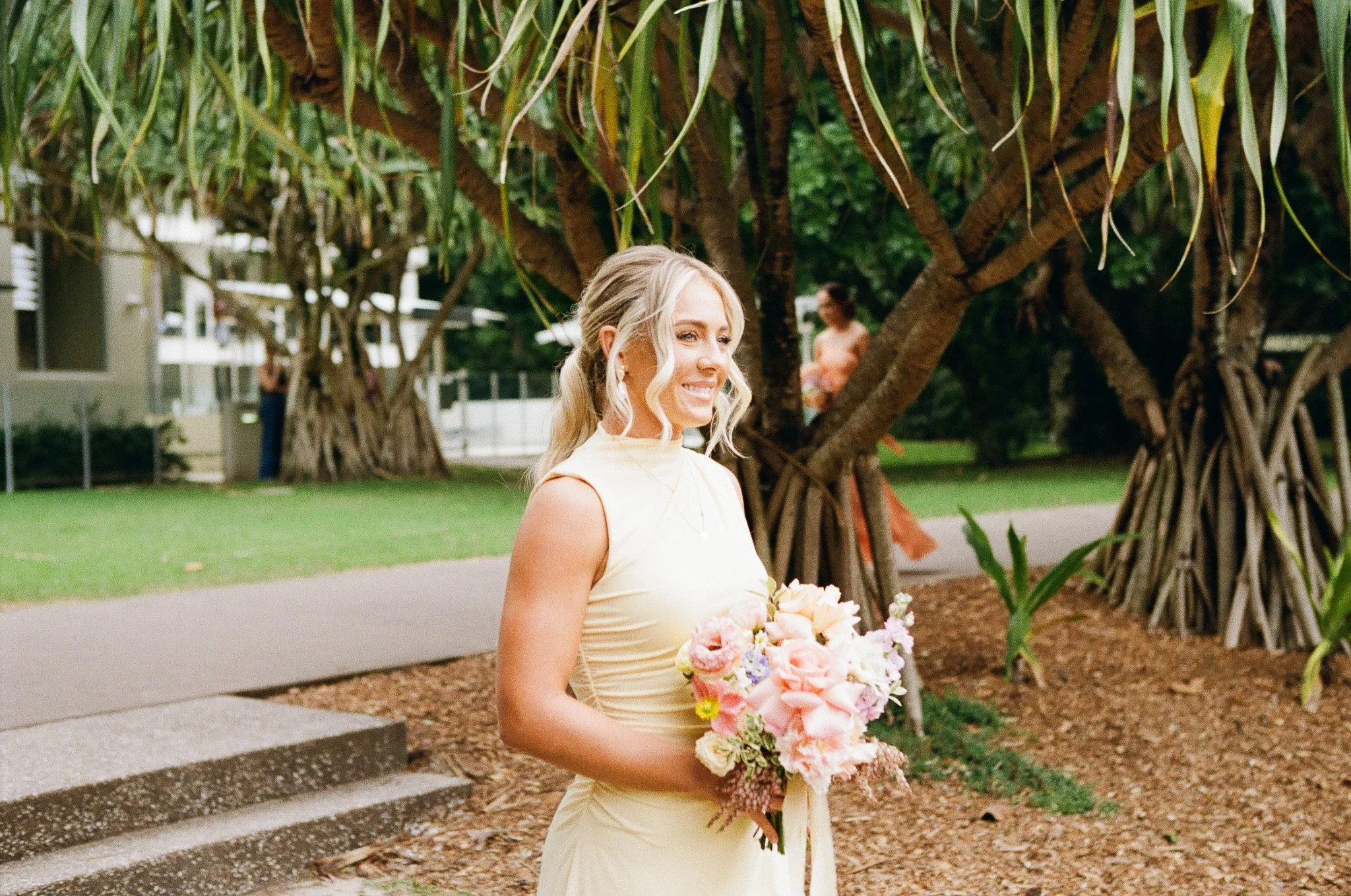 A woman in a cream-colored dress holding a bouquet of pink and white flowers, standing outdoors near large trees with thick roots, smiling.