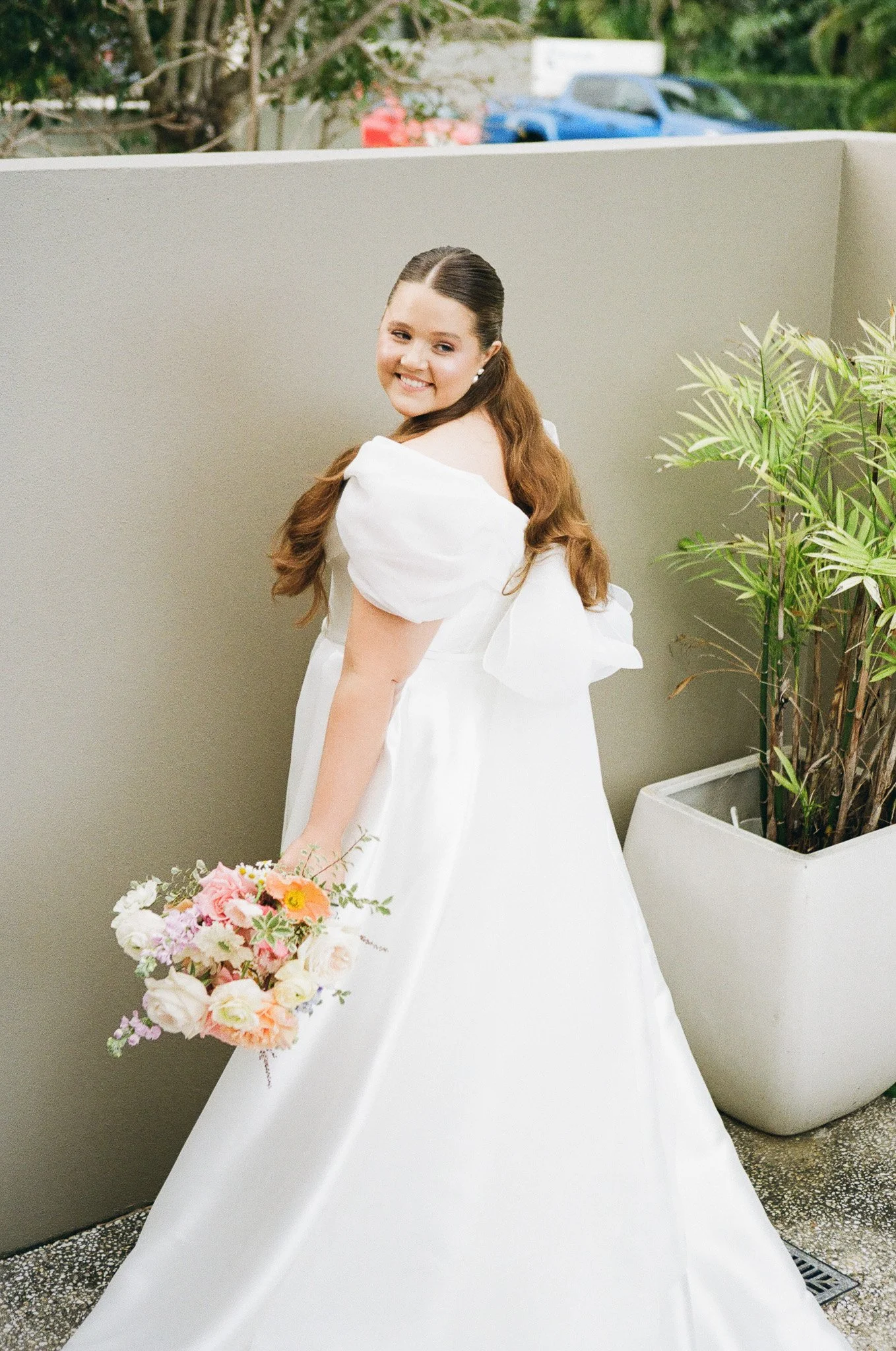 A young woman in a white wedding gown holding a bouquet of pink, white, and orange flowers, standing outdoors near a gray wall and green plants.
