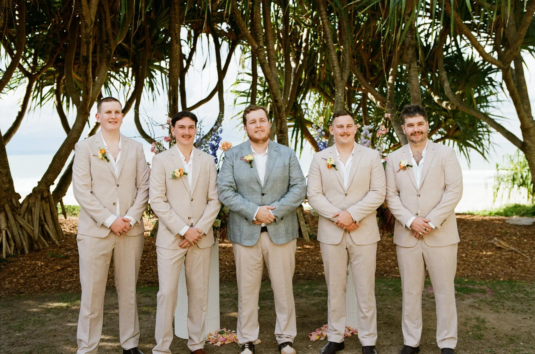 Groom and groomsmen standing outdoors under a large tree, dressed in suits with boutonnières, at a wedding ceremony.