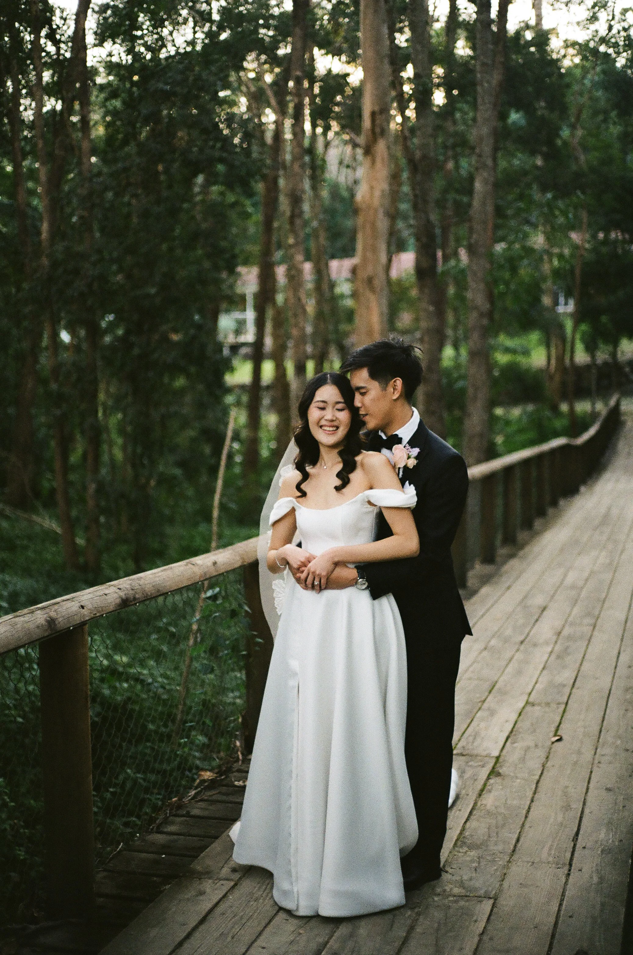 A bride and groom standing on a wooden bridge in a forest, embracing and smiling happily.