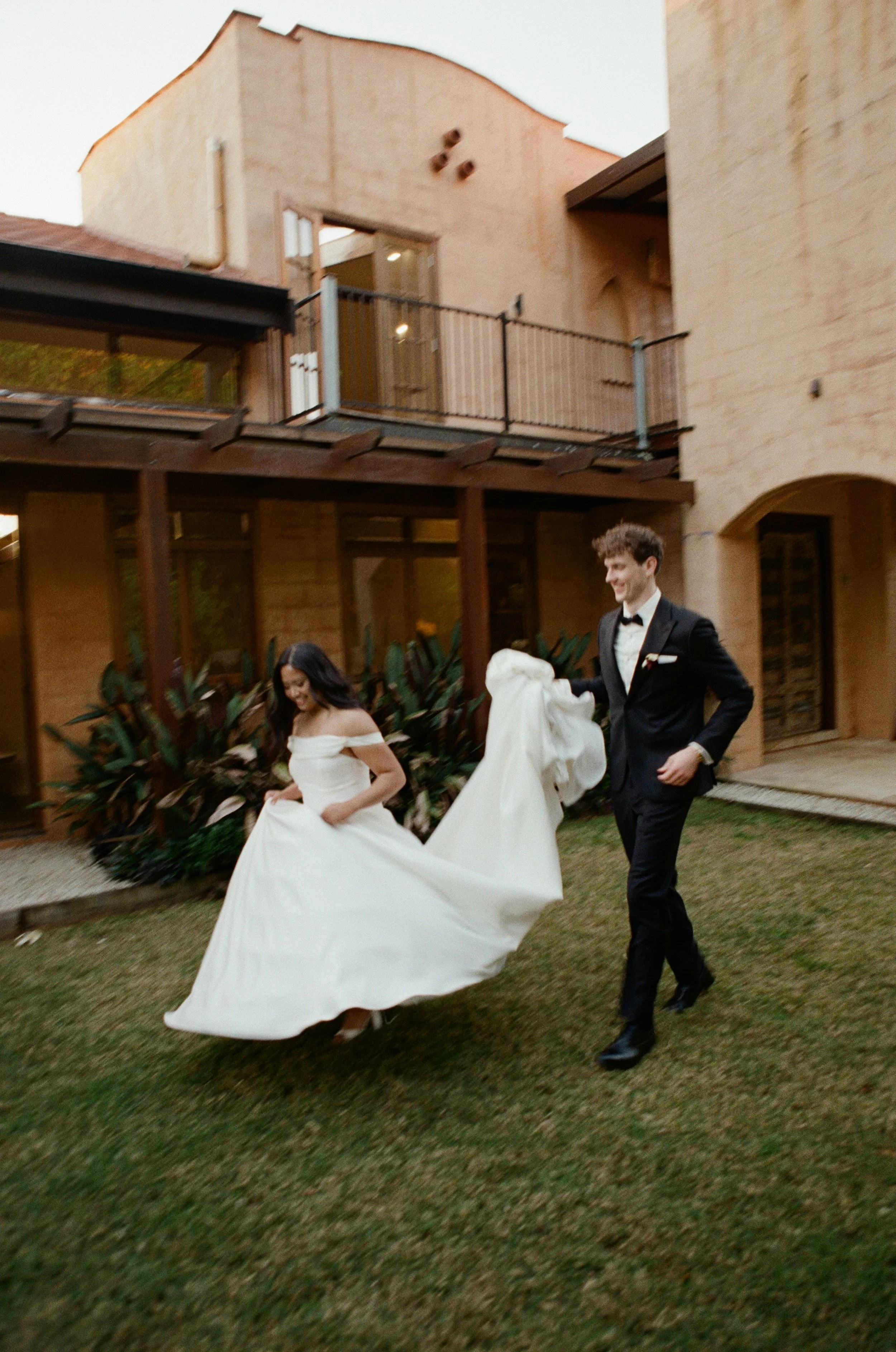 A bride and groom walking on a lawn, smiling; the bride is wearing a white off-shoulder wedding dress and holding her skirt, the groom is in a black tuxedo with a bow tie, holding the bride's train.