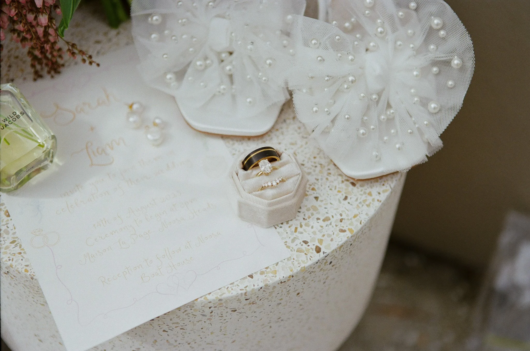 Wedding rings in a white velvet ring box, surrounding decor including pearl-embellished fabric, flowers, and an invitation on a terrazzo table.