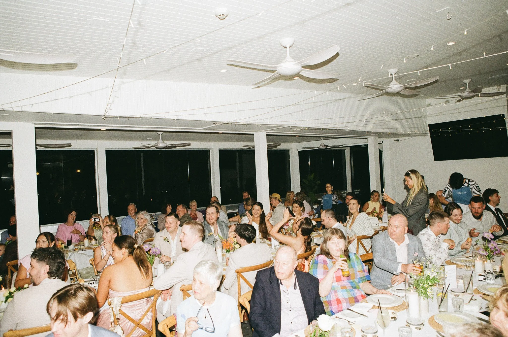 Guests seated at round tables with white tablecloths, floral centerpieces, and plates, attending a social event or dinner in a brightly lit room with large windows, ceiling fans, and a flat screen TV on the wall.