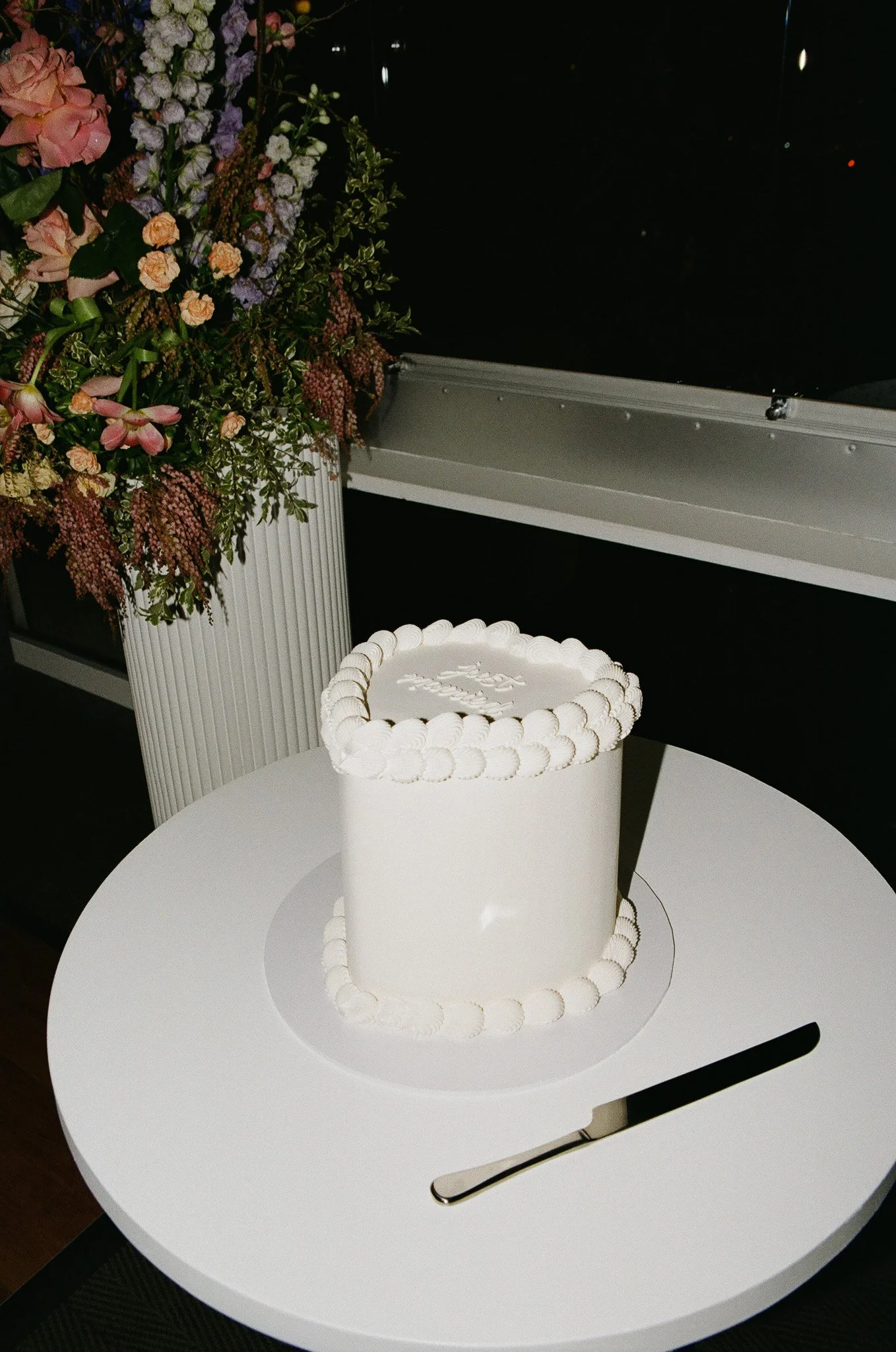 White round cake with white piped decoration on top and around the base, placed on a white circular base on a white table, with a flower arrangement in a tall white vase nearby.