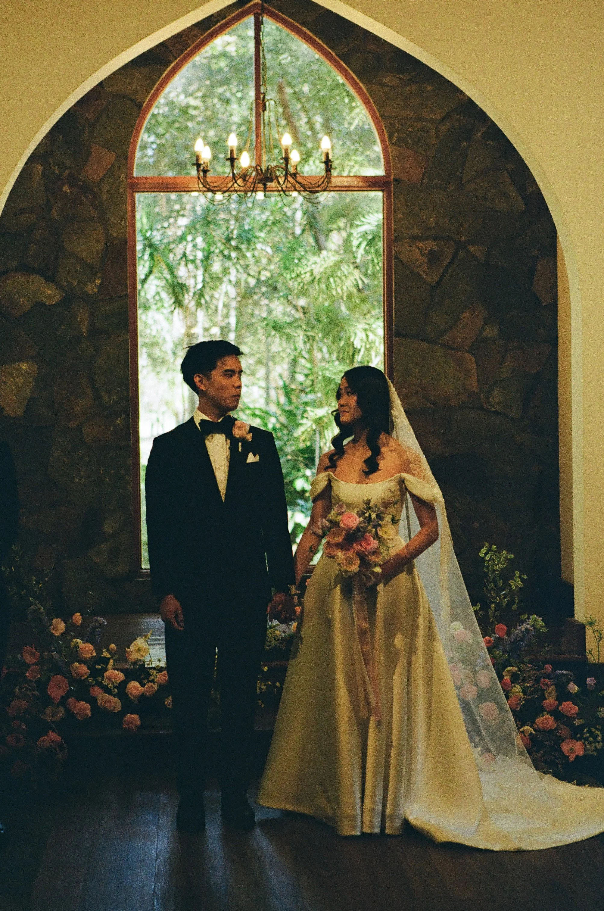 A couple dressed in wedding attire, holding hands, standing inside a church with a large window overlooking lush greenery outside, a chandelier hanging from the ceiling, and a floral arrangement at their feet.
