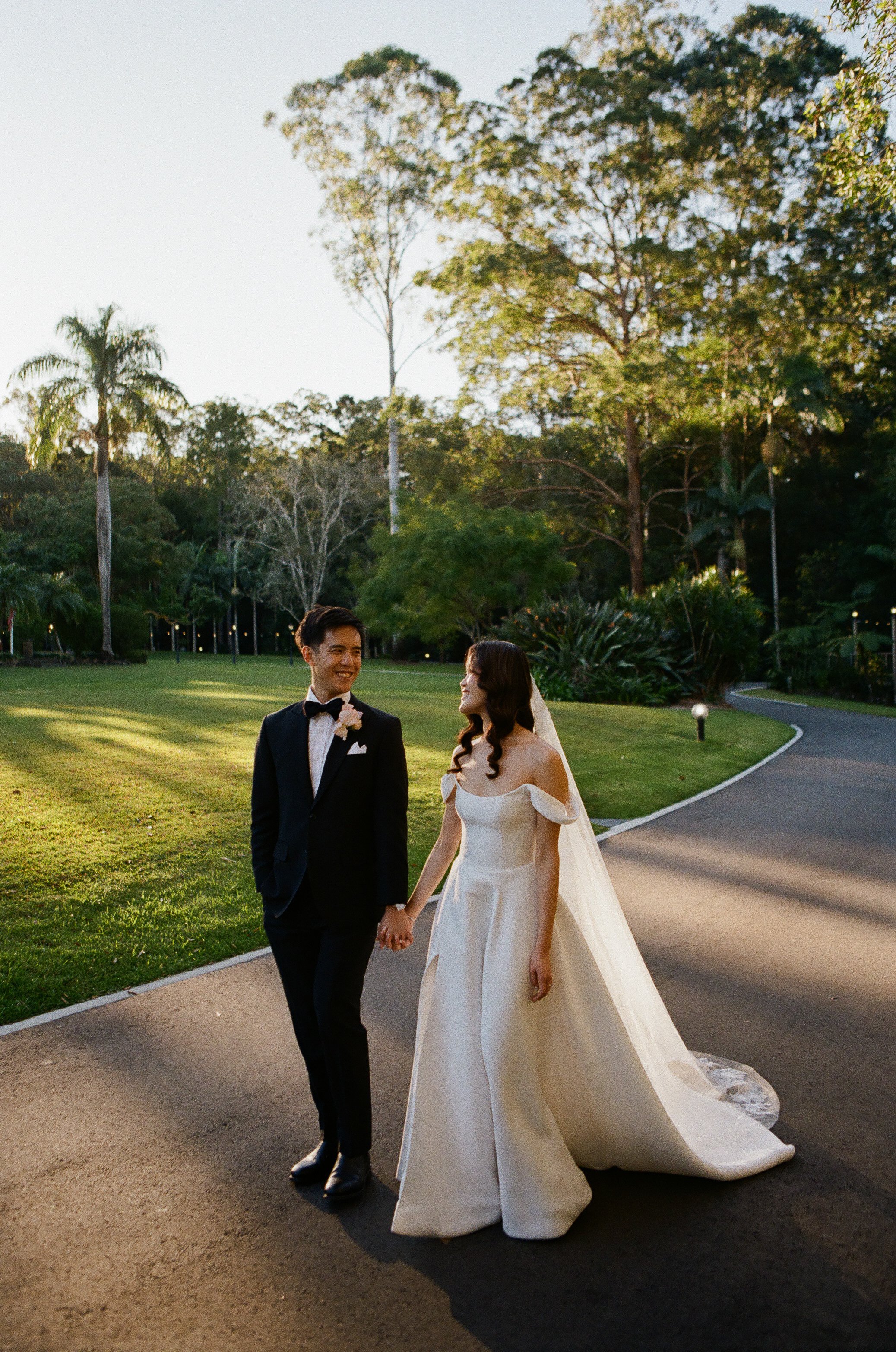 A bride and groom walking hand in hand outdoors during sunset, surrounded by trees and greenery.