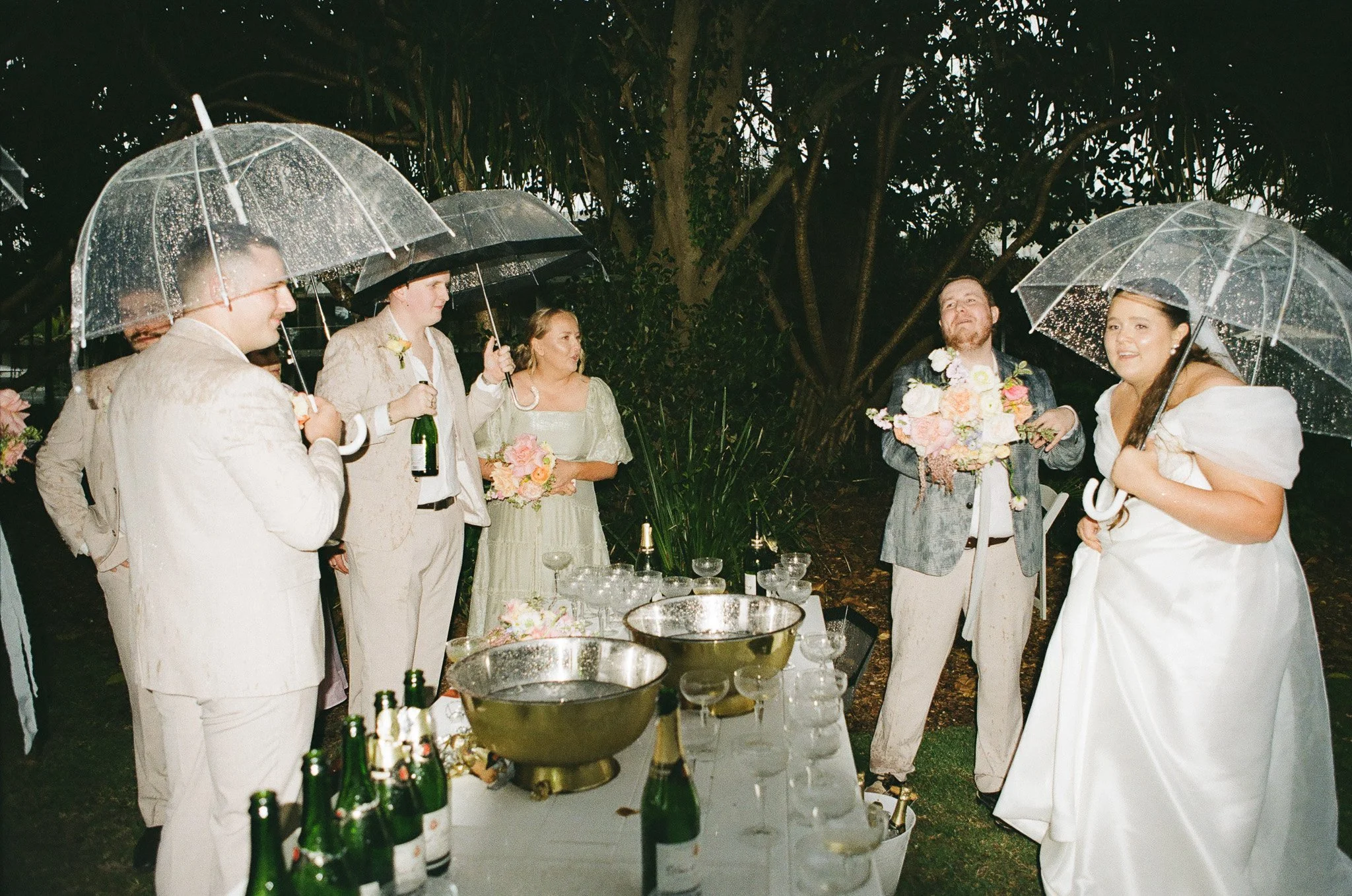 Wedding celebration outdoors in the rain with guests holding umbrellas, a table with champagne bottles and glasses, and a bride and groom holding bouquets.