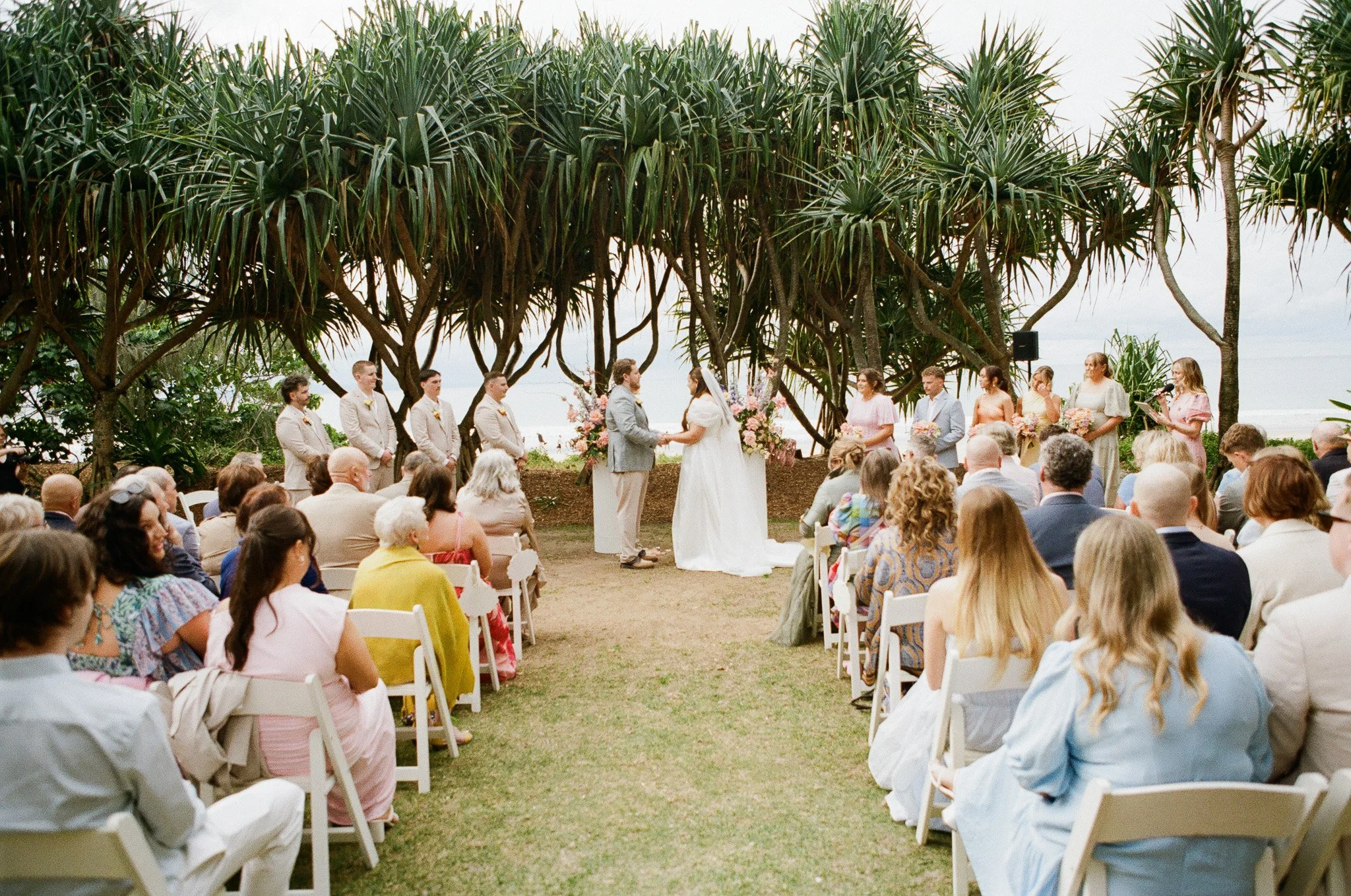 Outdoor wedding ceremony taking place under large trees with guests seated on white chairs, couple exchanging vows, floral arrangements, and bridesmaids and groomsmen standing nearby.