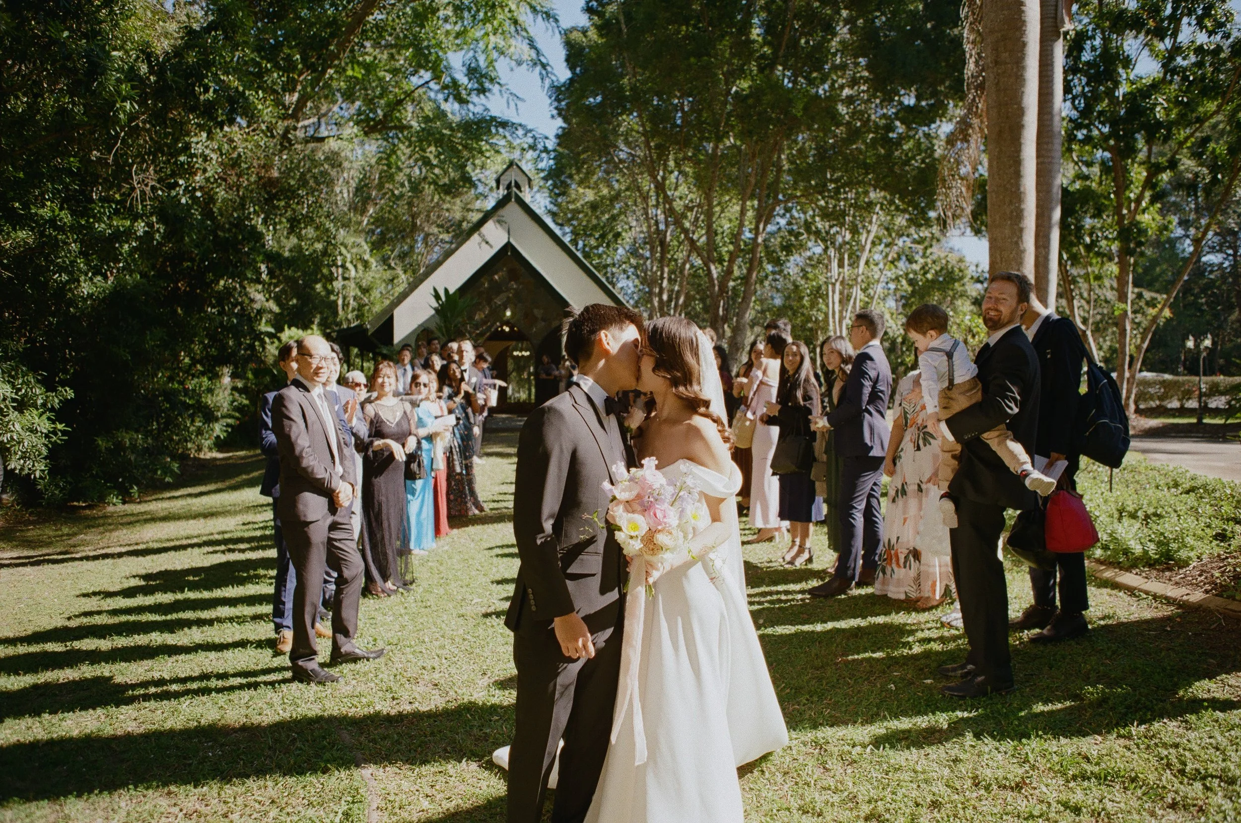 A bride and groom kissing during their outdoor wedding ceremony, with guests standing nearby on a sunny day in a park with trees and a chapel in the background.