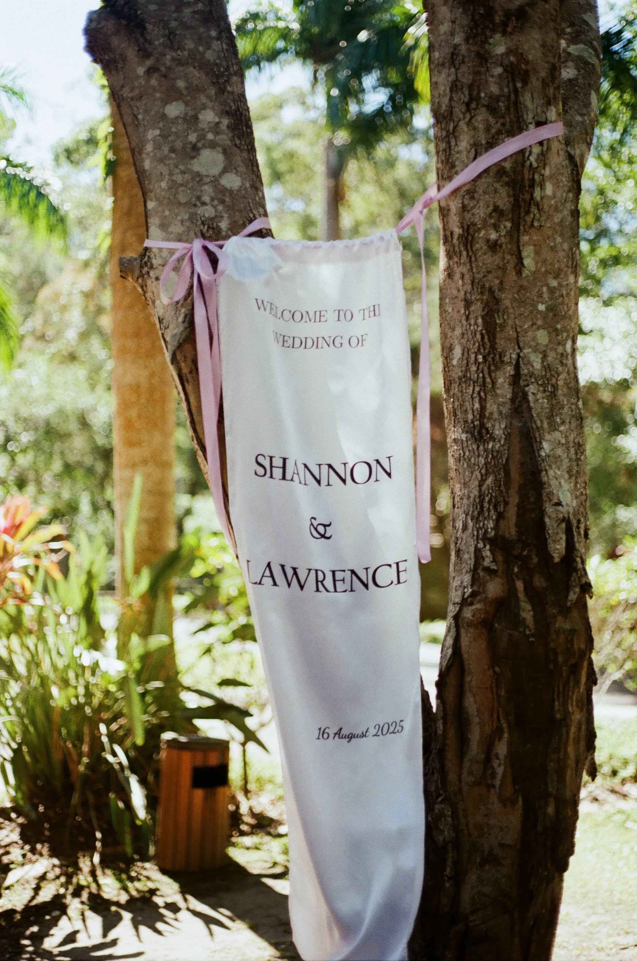 A wedding welcome sign hanging between two trees outdoors, reading "Welcome to the wedding of Shannon & Lawrence, 16 August 2025." The sign is decorated with pink ribbons in a garden setting.