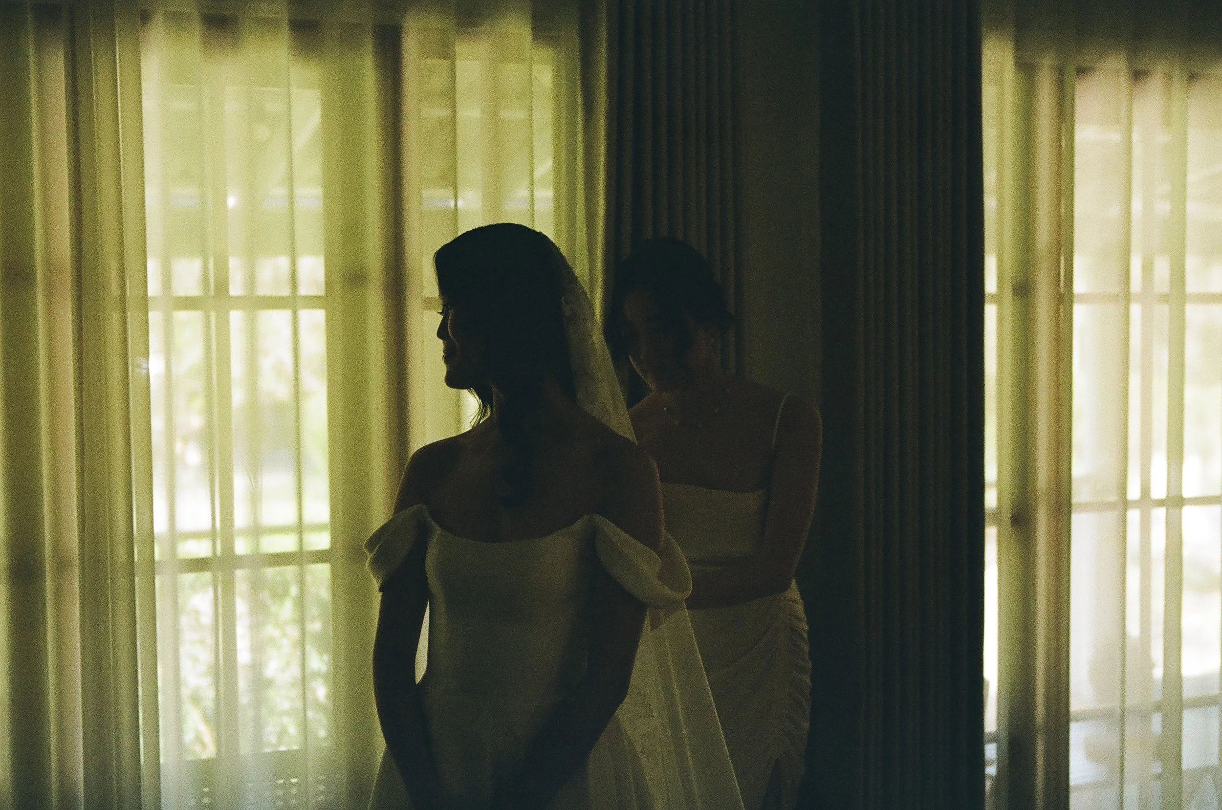 Silhouette of a woman in a wedding dress with off-the-shoulder sleeves, standing near a window with sheer curtains, and another woman in the background, both inside a room with dark vertical blinds.