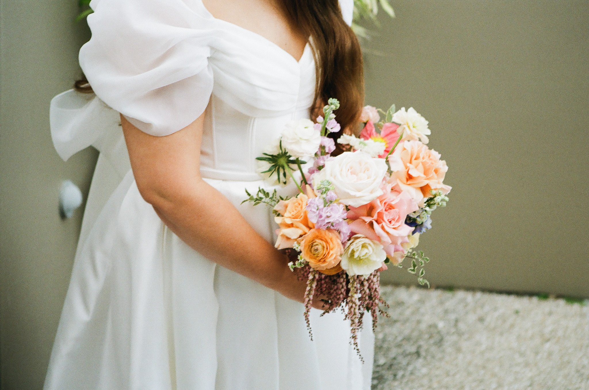 Close-up of a bride holding a bouquet of pink, white, and peach roses and other flowers, dressed in a white gown with puffed sleeves.