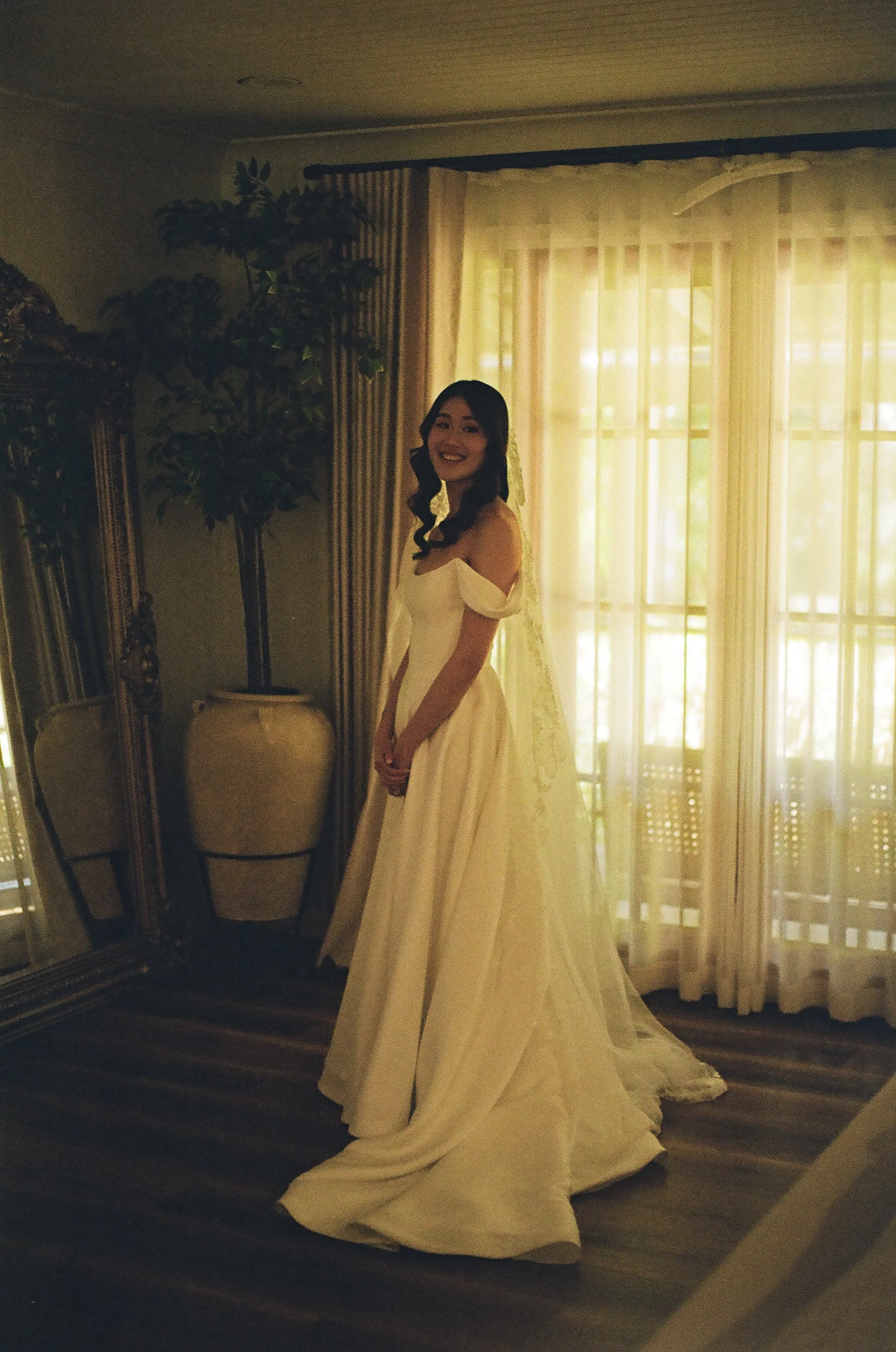 A woman in a wedding dress standing indoors near a window with curtains, smiling.