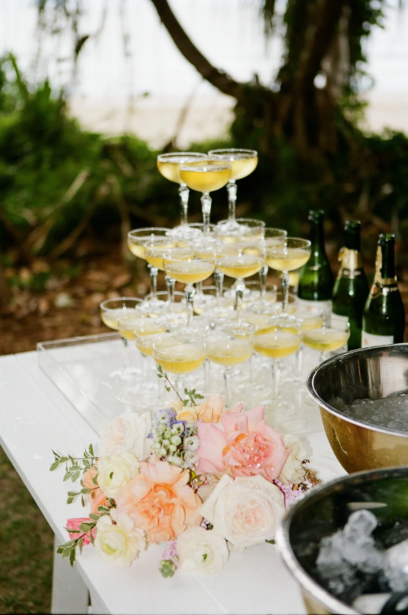 Several champagne glasses filled with a yellow drink arranged in a pyramid on a white table, with a floral centerpiece and bottles of champagne and ice buckets nearby outdoors.