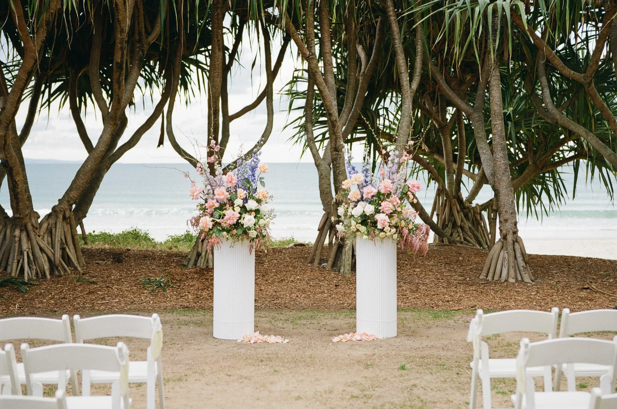 A beach setup with two white pedestals holding flower arrangements in front of trees, overlooking the ocean, with white chairs arranged around.
