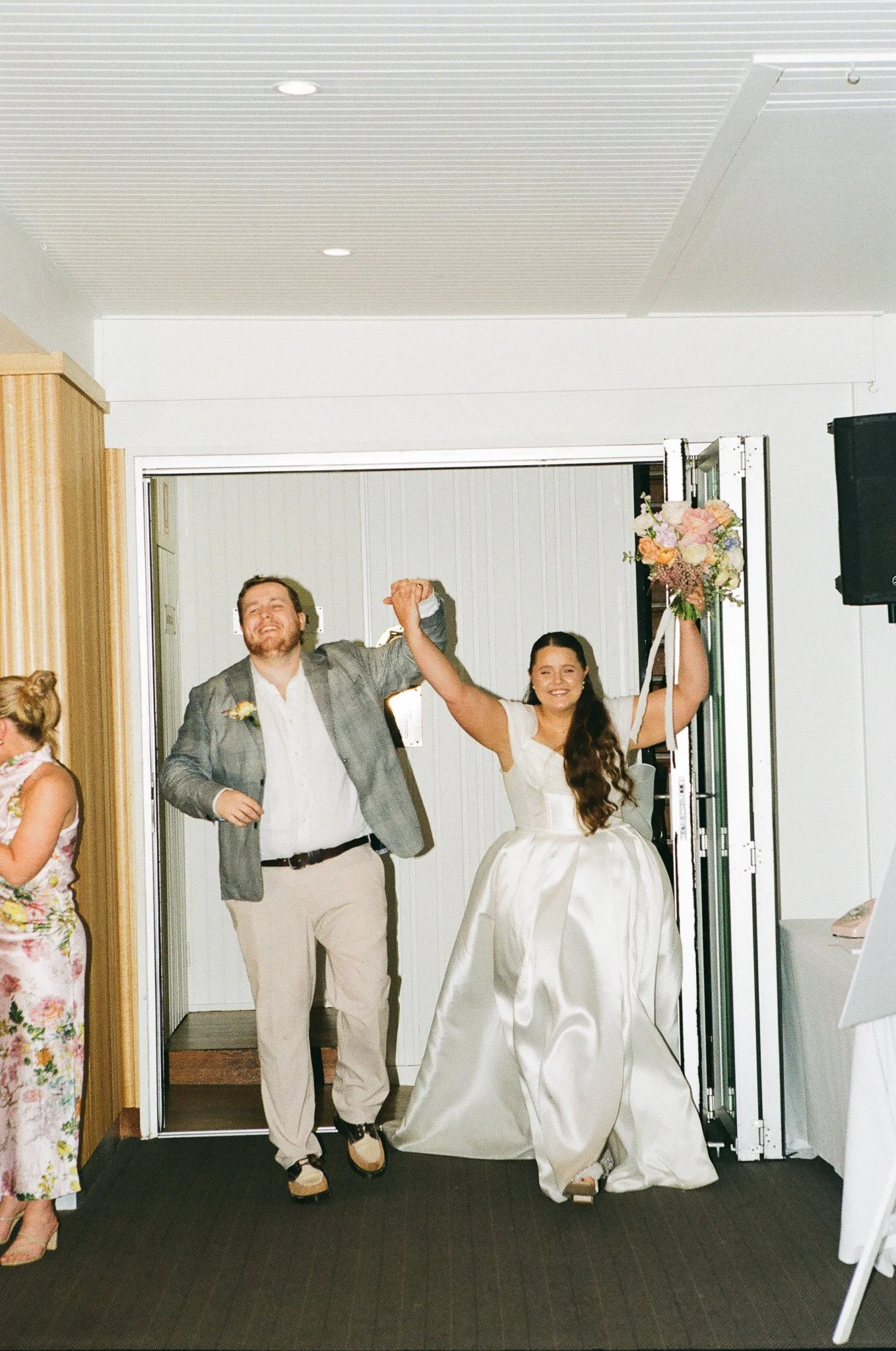 A newlywed couple, the bride in a white gown holding a floral bouquet, and the groom in a gray blazer and beige pants, smiling and holding hands as they exit a building.