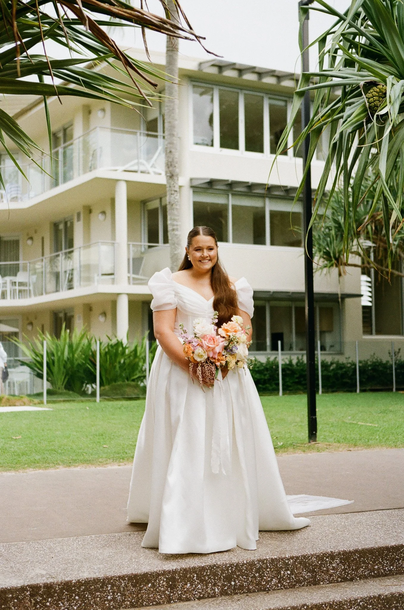 A bride in a white wedding dress holds a colorful bouquet of flowers and smiles outdoors in front of a modern building with balconies.