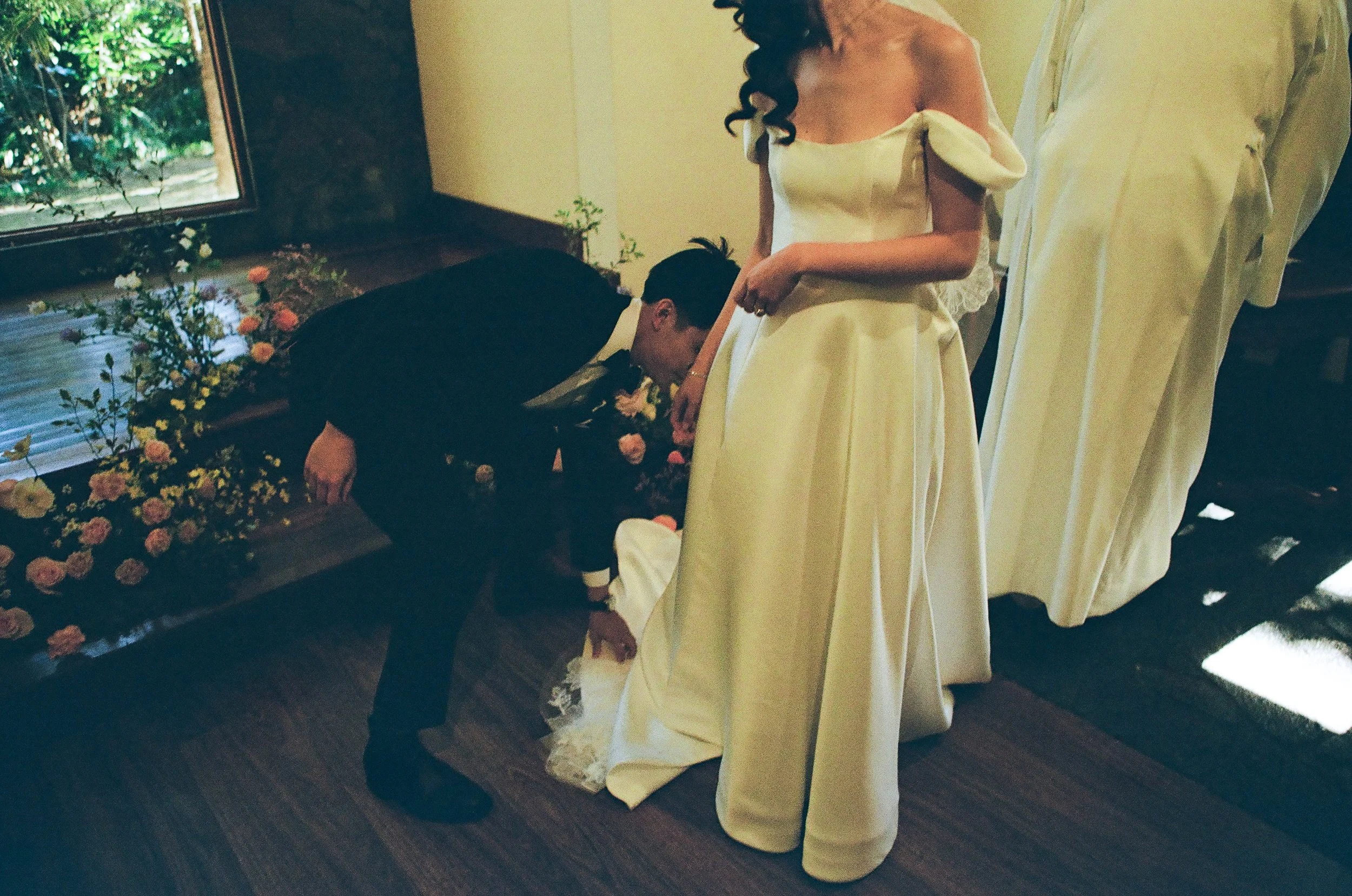 A bride in a white wedding dress stands while a person adjusts her dress, with another person in white standing nearby. The scene takes place indoors near a window with flowers outside.