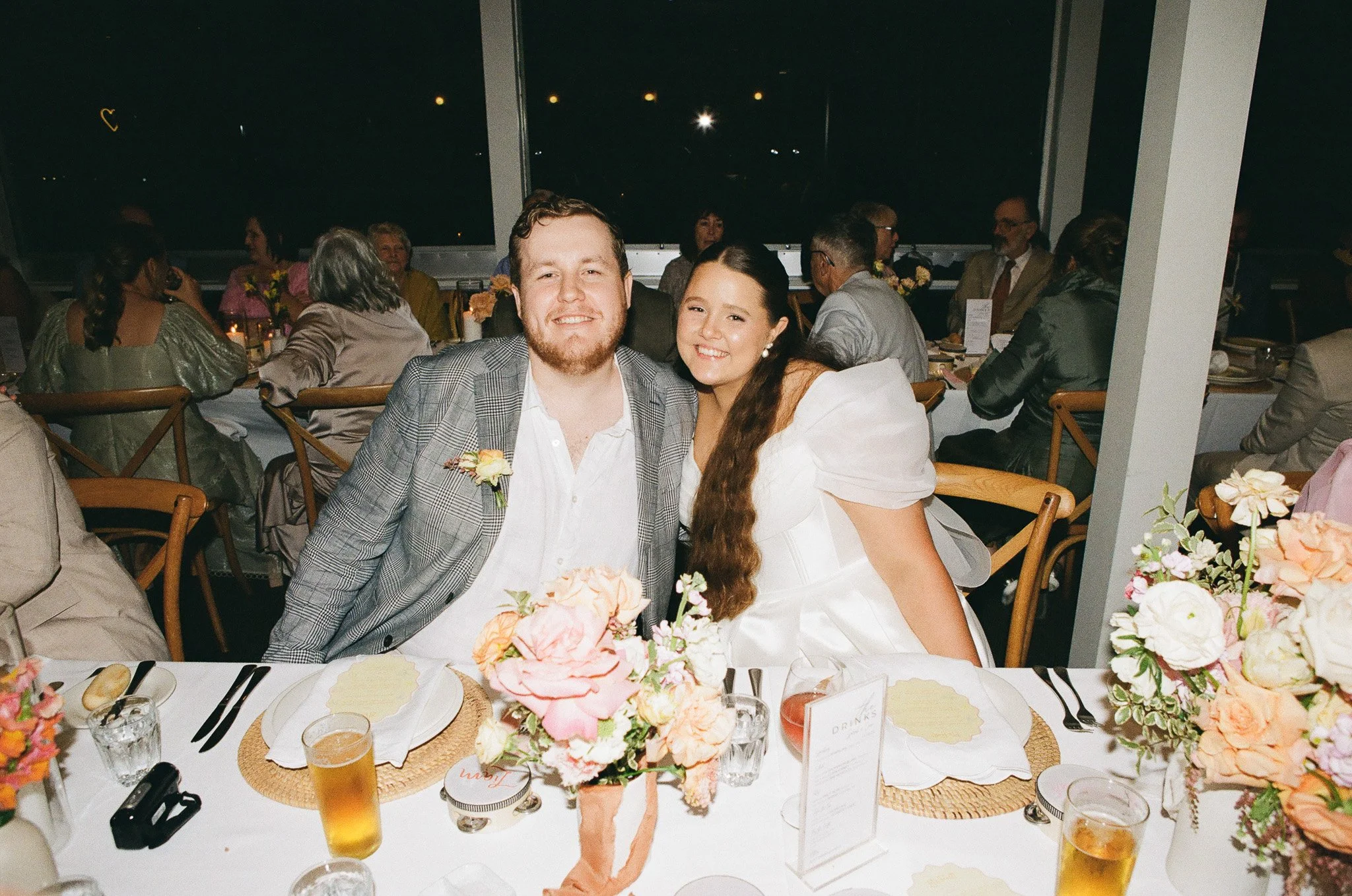 Young man and woman in wedding attire sitting at a decorated reception table, smiling at the camera, with multiple guests in the background at a nighttime event.