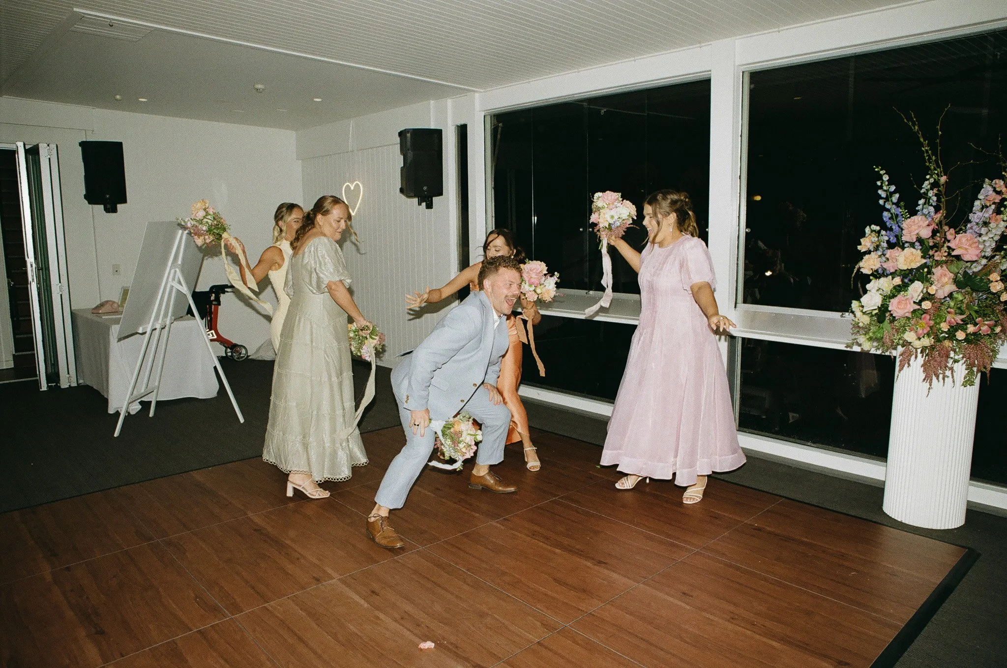 People dancing at a wedding reception, holding bouquets, in a decorated indoor venue.