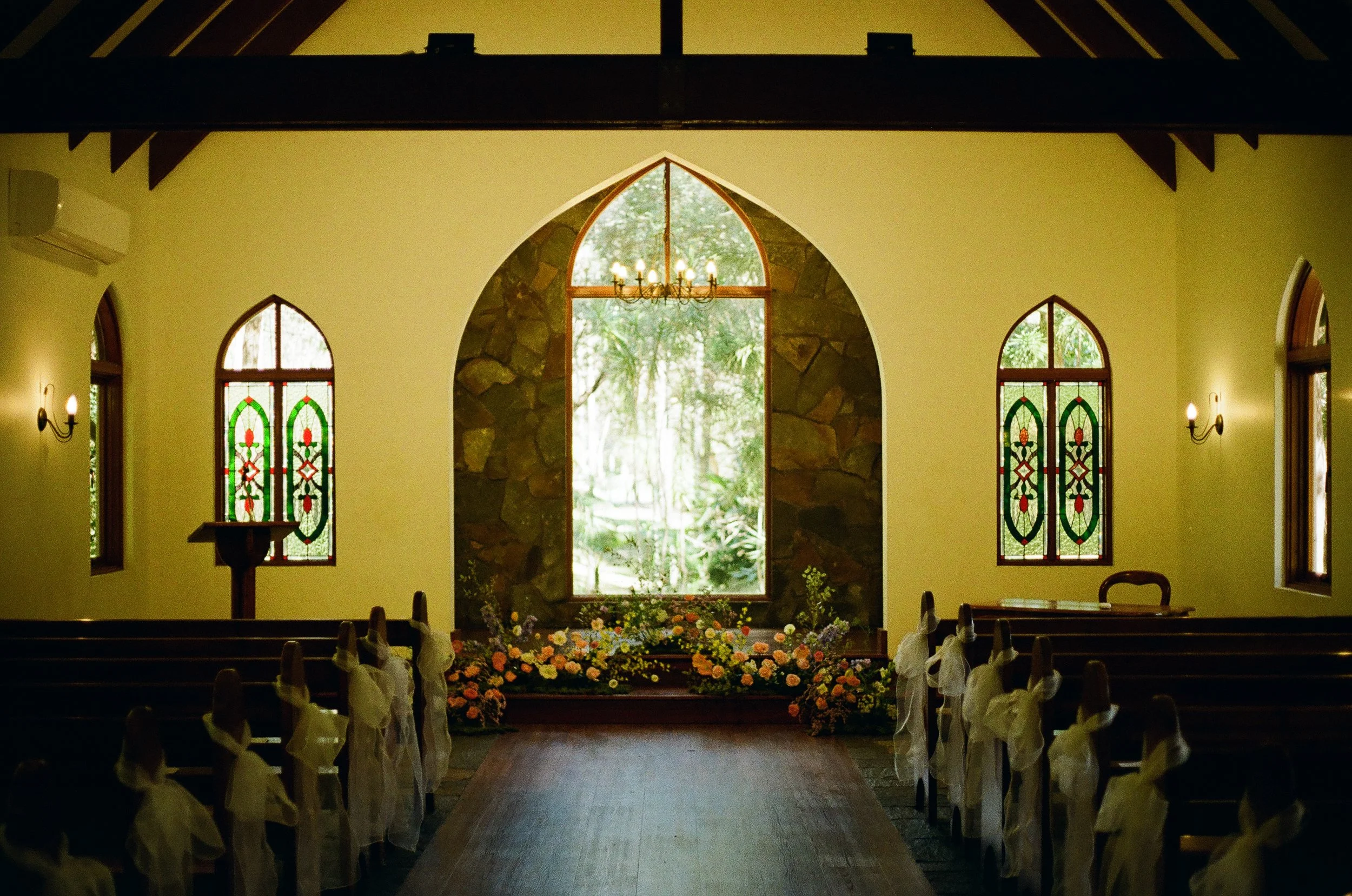 Interior of a small chapel decorated for a wedding, with pews adorned with white bows, stained glass windows, and a floral arrangement at the altar in front of large arched windows showing trees outside.