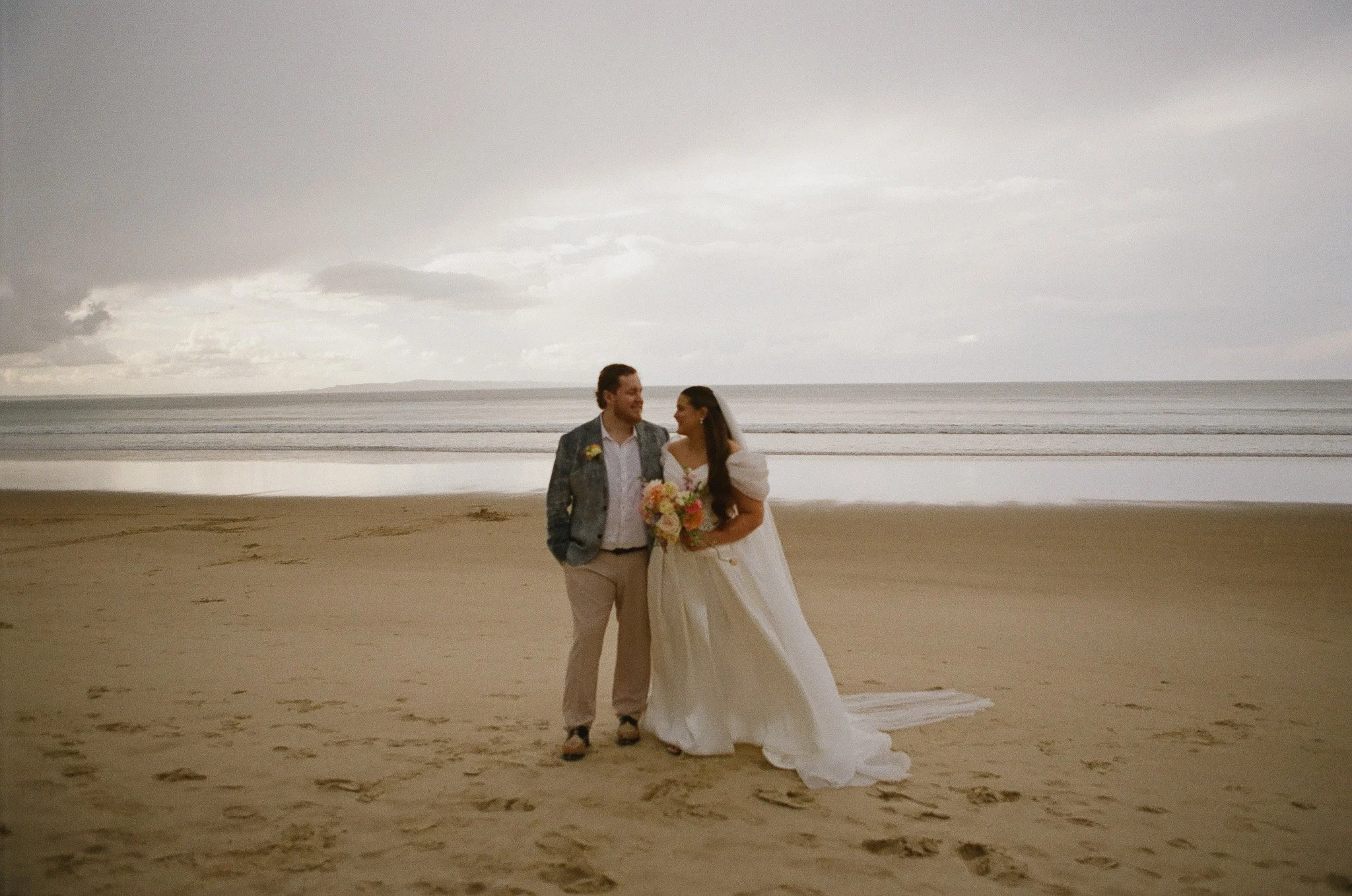 Bride and groom standing on the beach during their wedding, holding a bouquet of flowers, with ocean and cloudy sky in the background.