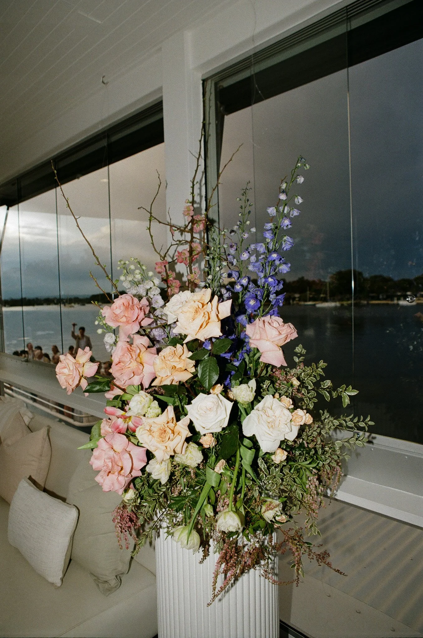A large floral arrangement of pink, white, and purple flowers in a white vase, placed on a table near a large window overlooking a body of water with a cloudy sky.