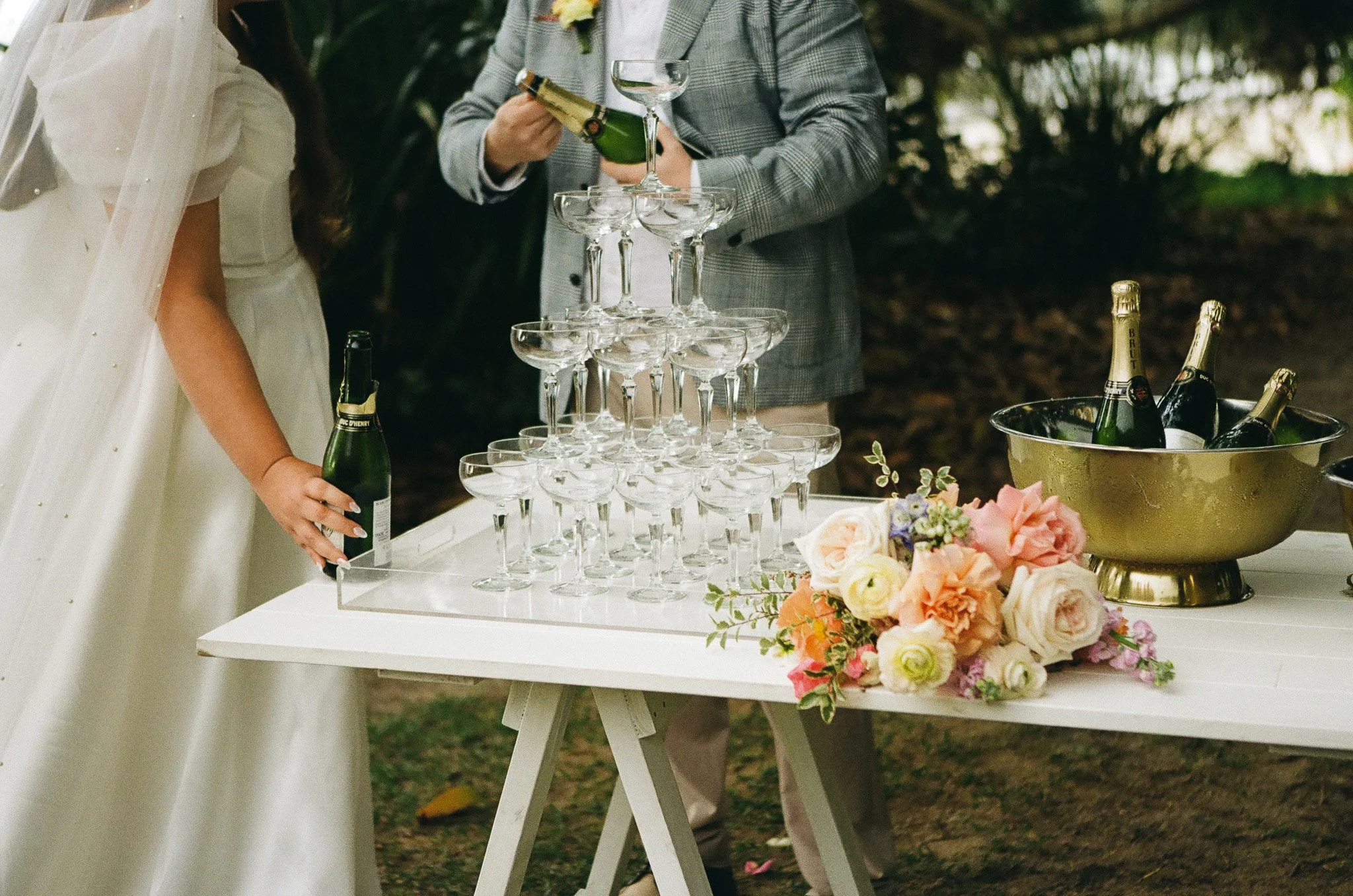 Wedding reception scene with a champagne bottle and glasses forming a pyramid on a white table, flowers, and a bucket of champagne bottles on an outdoor setting.
