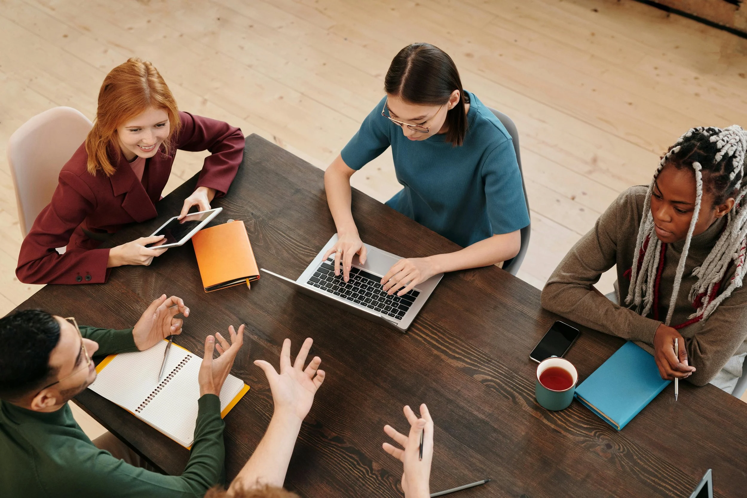 Group of diverse women sitting around a wooden table engaged in discussion, some taking notes, using laptops and tablets, with notebooks, pens, a smartphone, and a mug of tea visible.