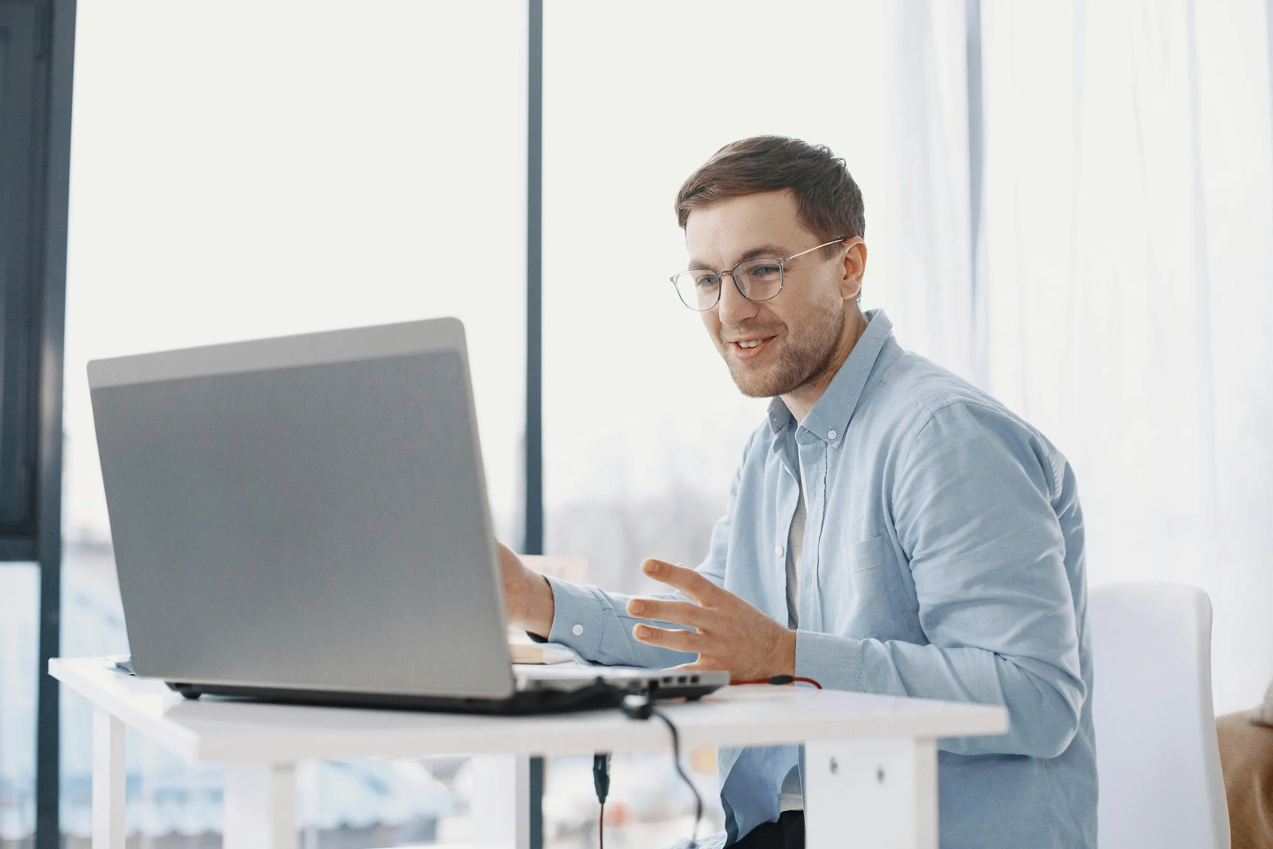A young man with brown hair and glasses, wearing a light blue shirt, sitting at a white table working on a laptop, smiling, in a bright room with large windows.