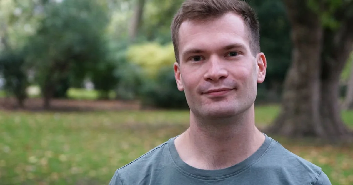 A young man with short brown hair and fair skin, wearing a gray t-shirt, standing outdoors in a park with green trees and grass in the background.