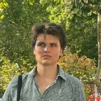 A man with short dark hair wearing a gray patterned shirt outdoors among trees and greenery.
