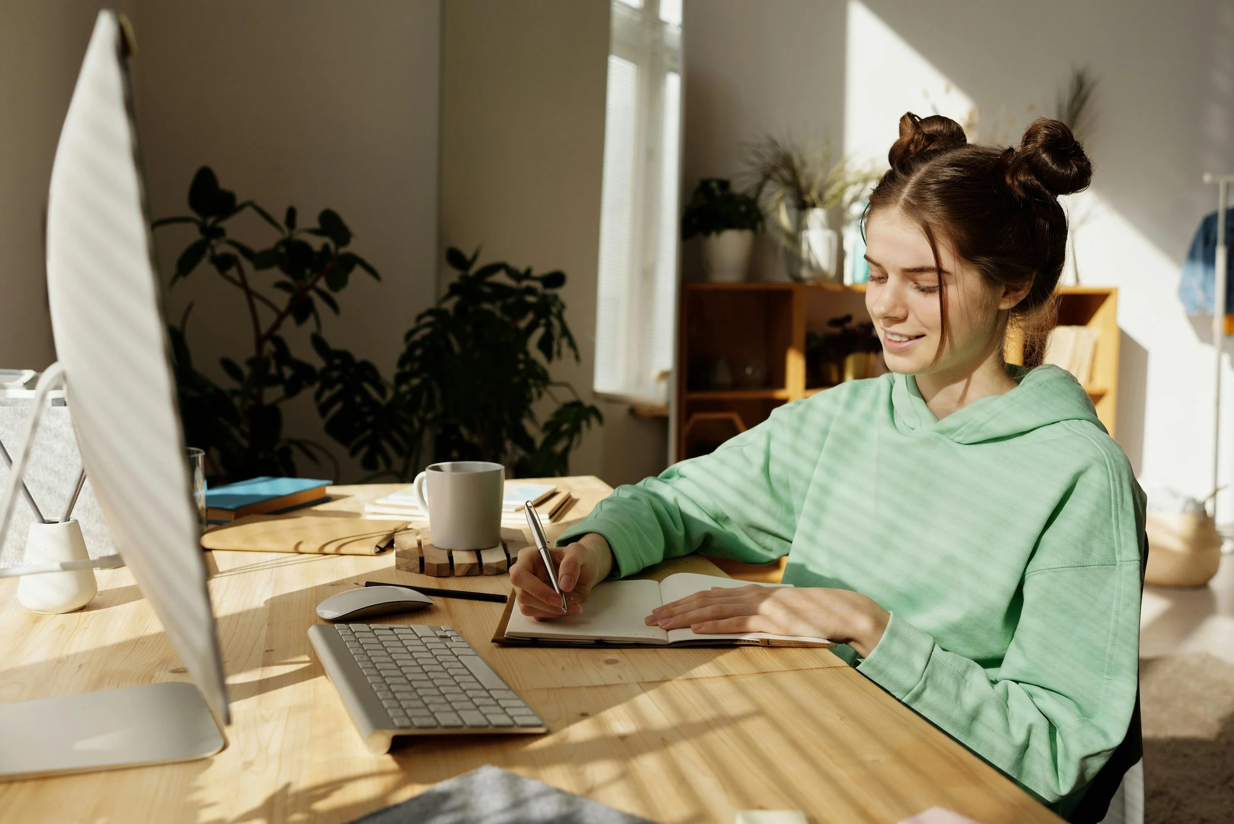 A young woman with brown hair styled in two buns, wearing a light green hoodie, sitting at a wooden desk in a bright room, writing in a notebook. There is a computer, a mug, and some notebooks on the desk, with plants and a wooden cabinet in the background.