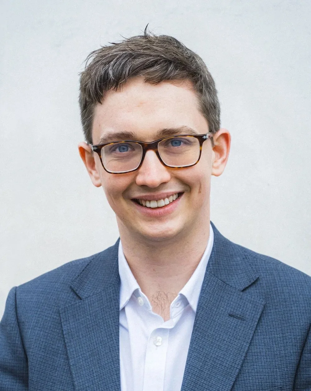 A young man with short brown hair, glasses, and a bright smile, wearing a blue blazer and white shirt, standing against a plain light background.