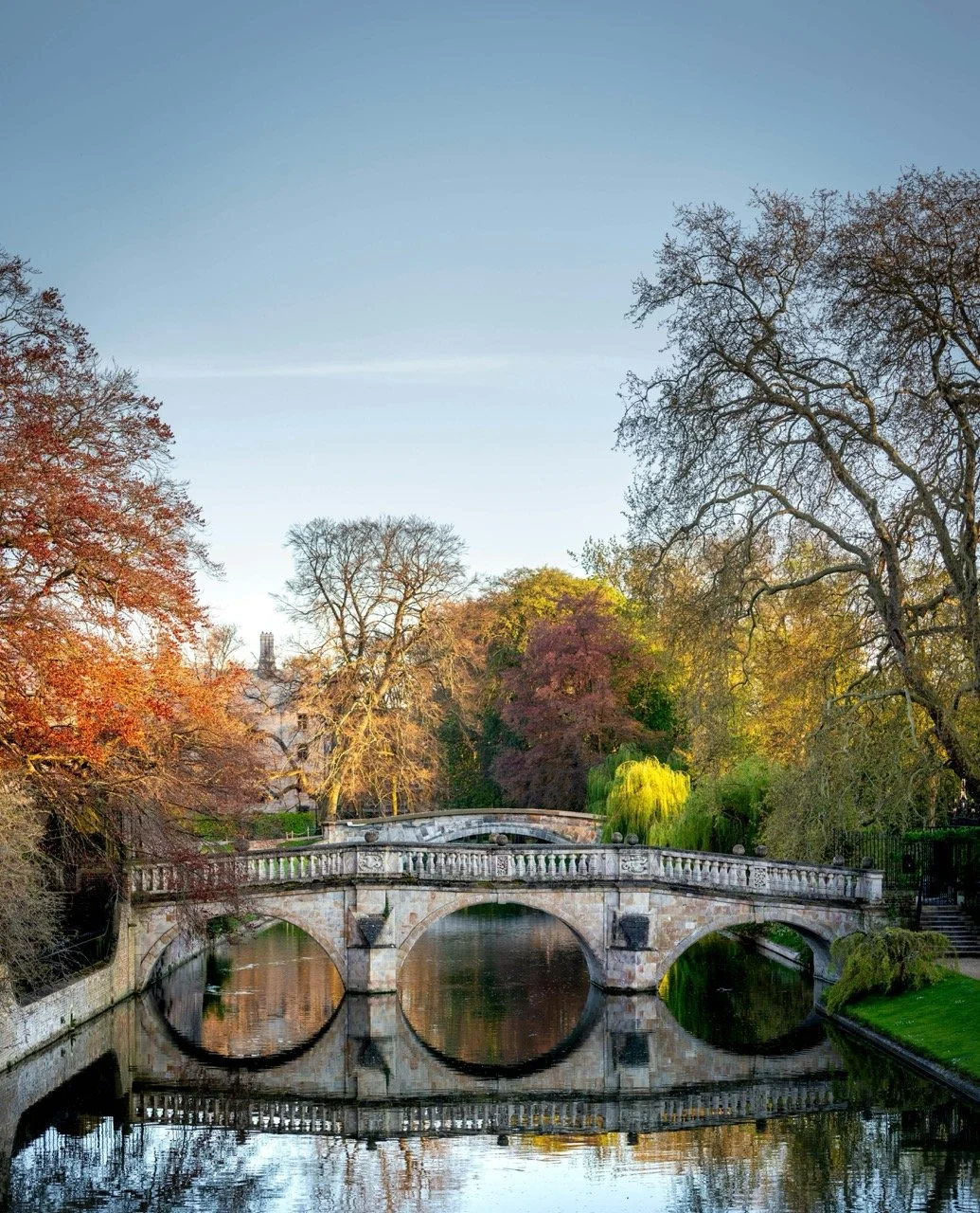 The Bridge of Sighs in Cambridge - a stone bridge over a calm river surrounded by trees with colorful autumn foliage and a clear sky.