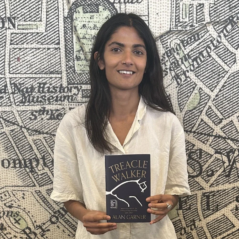 A woman with long dark hair and a nose ring holding a book titled 'Treacle Walker' by Alan Garner, standing in front of a black-and-white illustrated map background.