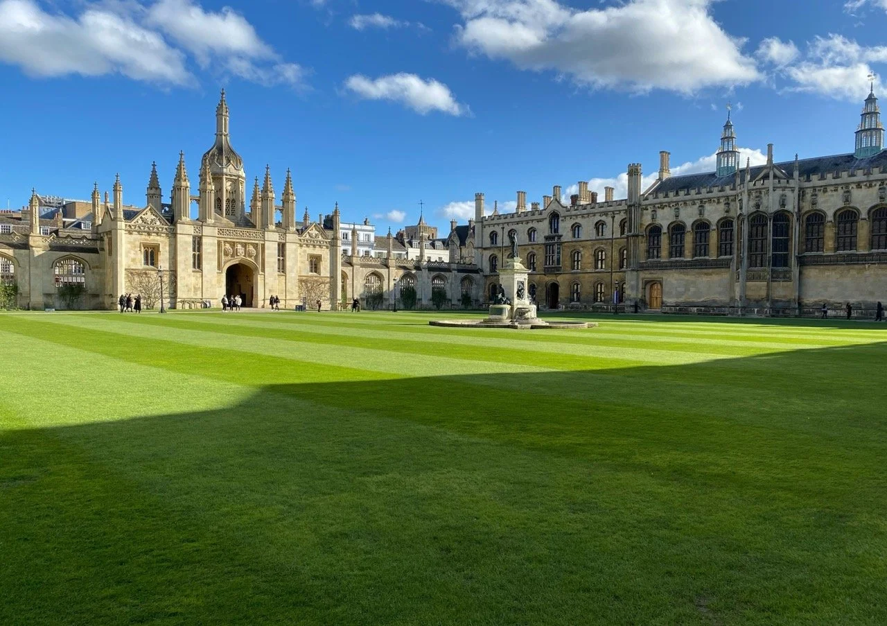 View of a historic building and quad at Trinity College, Cambridge, with gothic architecture, a well-maintained grassy courtyard, and a fountain in front under a partly cloudy sky.