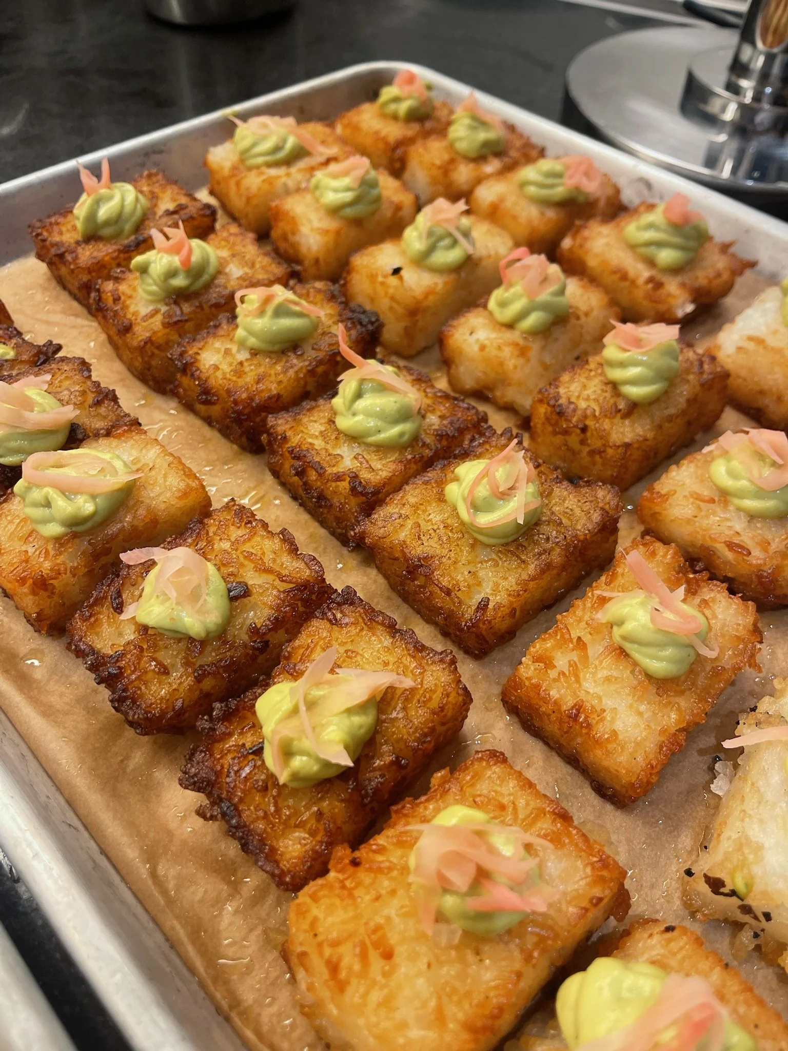 Crispy fried fish pieces topped with green wasabi mayo and pink pickled ginger on a baking sheet.