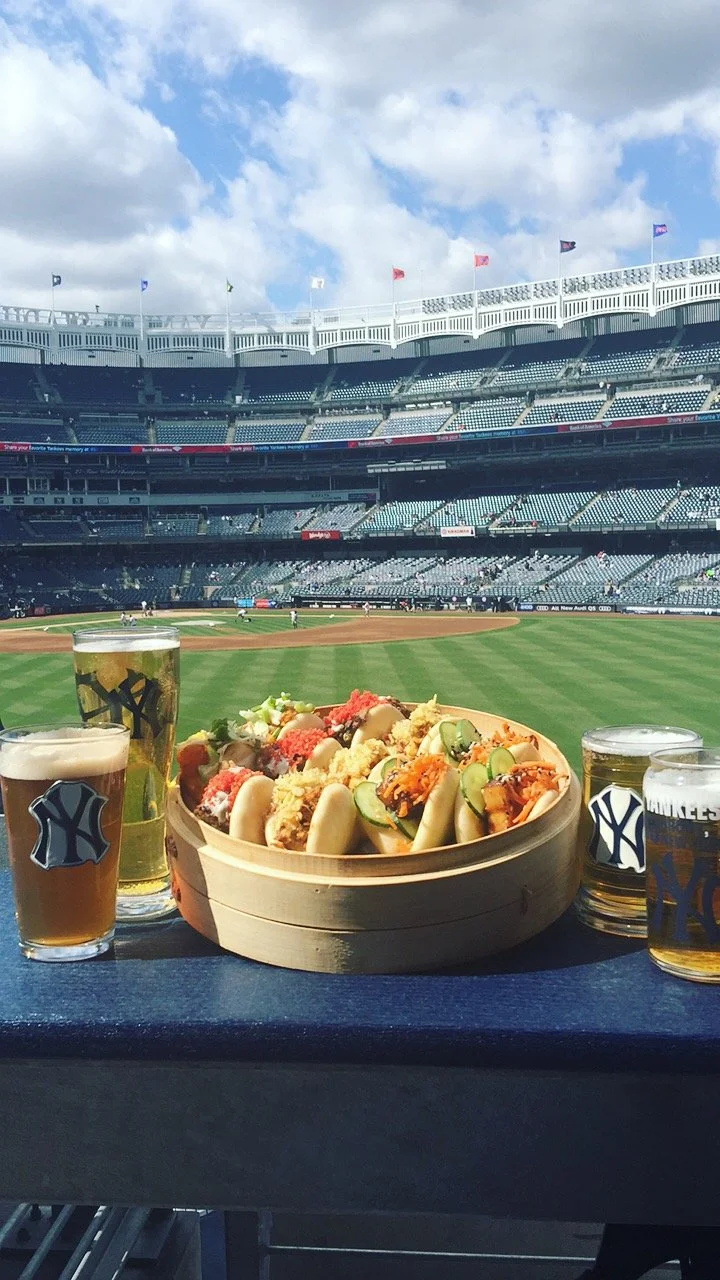 View of a baseball stadium with food and drinks on a table in the foreground, including four glasses of beer and a tray of hot dogs topped with various condiments. The field is visible in the background under a partly cloudy sky.