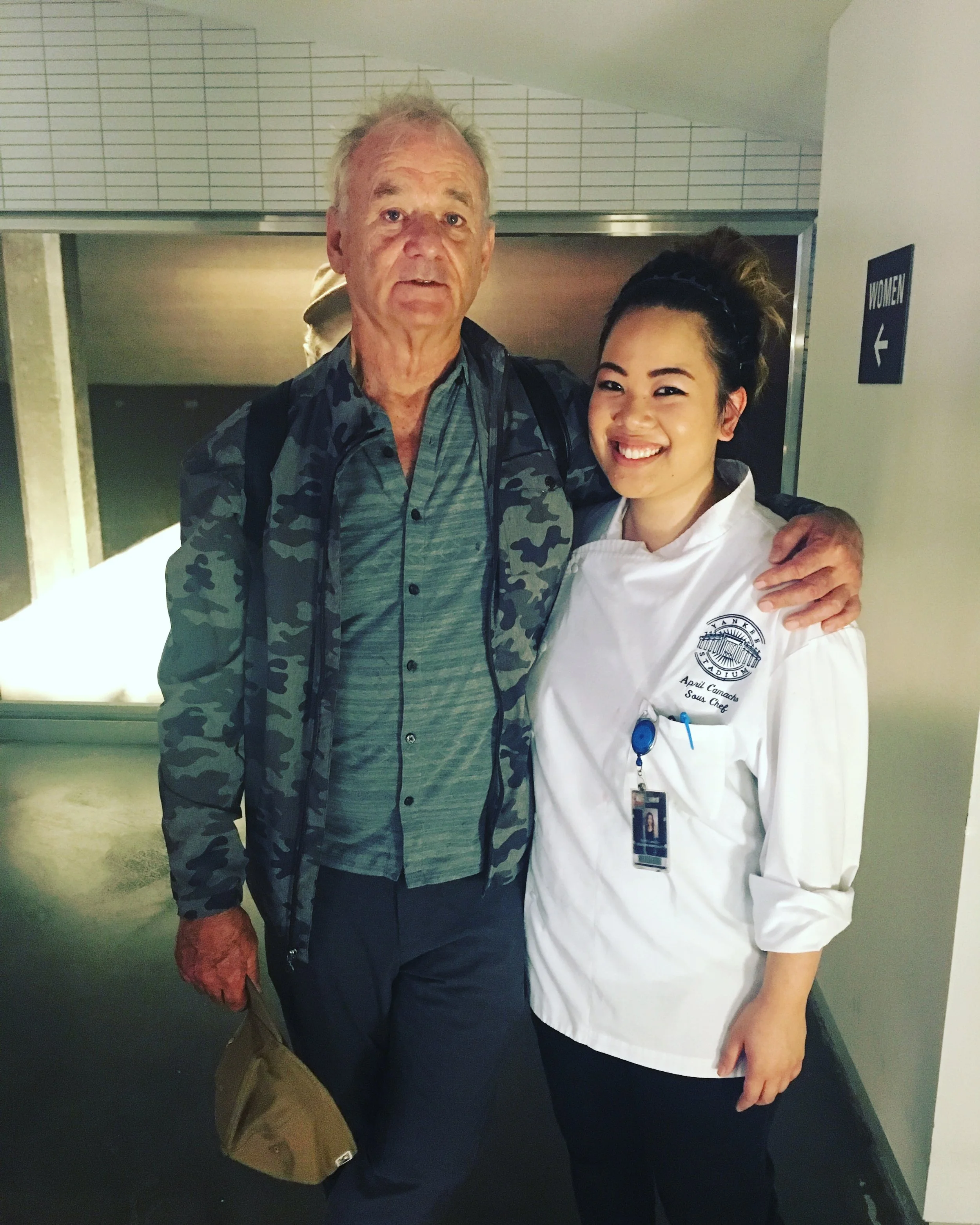 An elderly man and a young woman in a chef uniform are standing together and smiling, with the man’s arm around her shoulders, in a corridor near a women’s restroom sign.