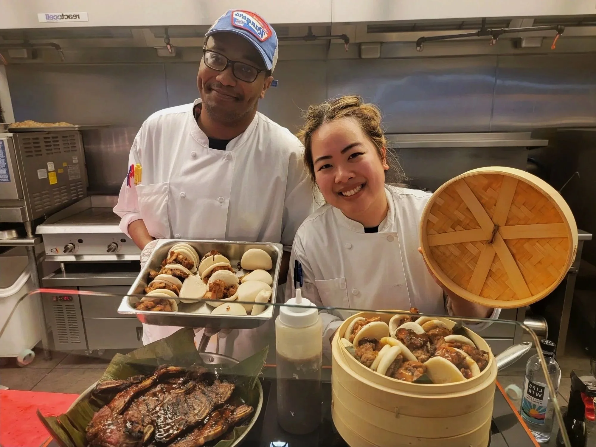 Two chefs in white uniforms standing in a kitchen with Asian dishes, including buns and grilled food, and a bamboo steamer. One chef holds a tray of buns, and the other holds a bamboo steamer